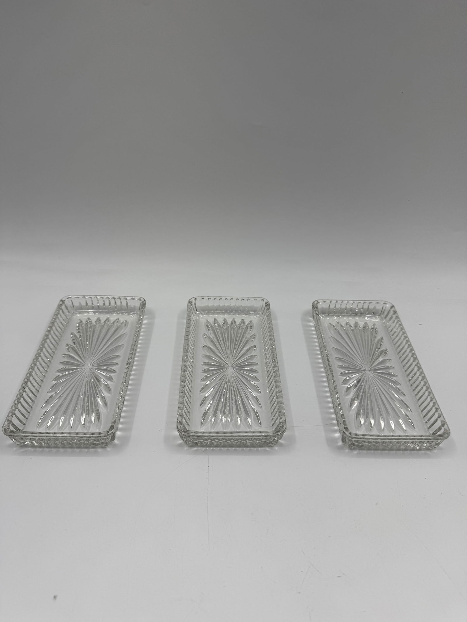 Set of Three Clear Glass Trays: Set of three clear glass trays, each approximately 7.5 x 4 inches. Features a starburst design on the base and a textured rim. Ideal for serving or display.