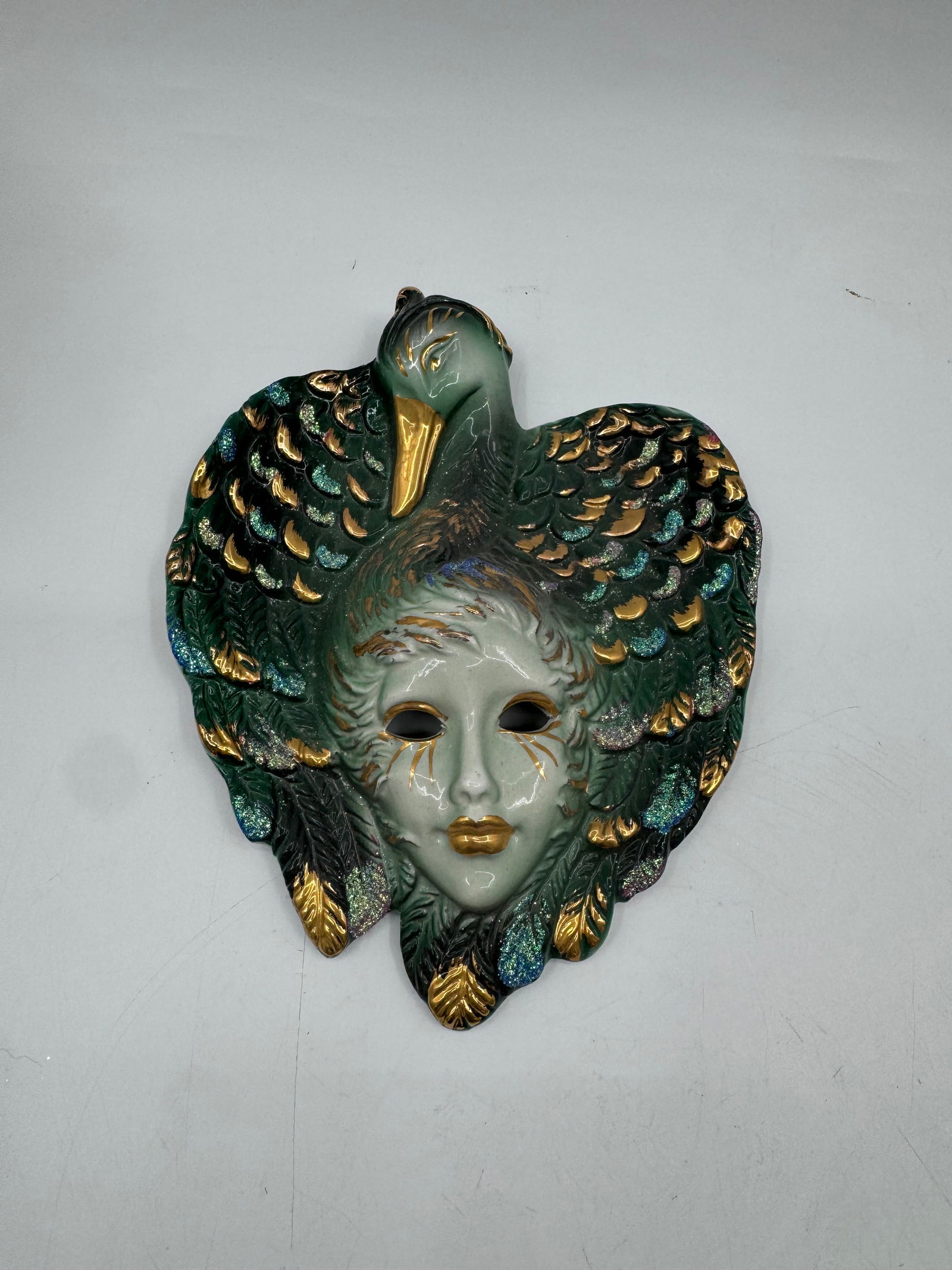 Venetian Style Wall Mask with Bird: Venetian style wall mask featuring a stylized face with a bird figure above. Green, gold, and white coloration. Measures approximately 8 x 6 inches.