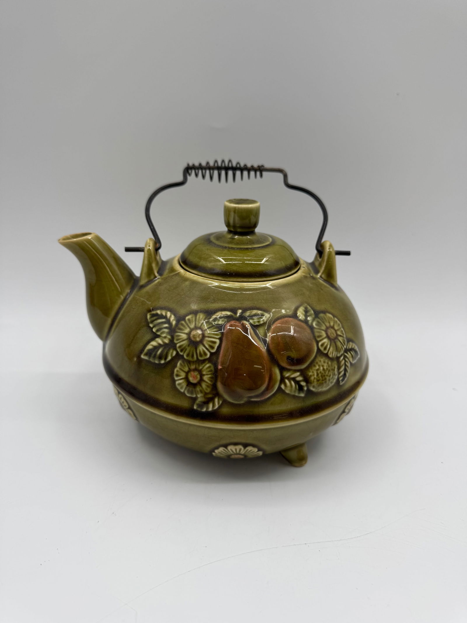 Vintage Olive Green Ceramic Teapot with Fruit Design: Vintage olive green ceramic teapot featuring a raised fruit and floral design. The teapot has a metal handle and a matching lid. Dimensions: 6" H x 8" L x 7" W.