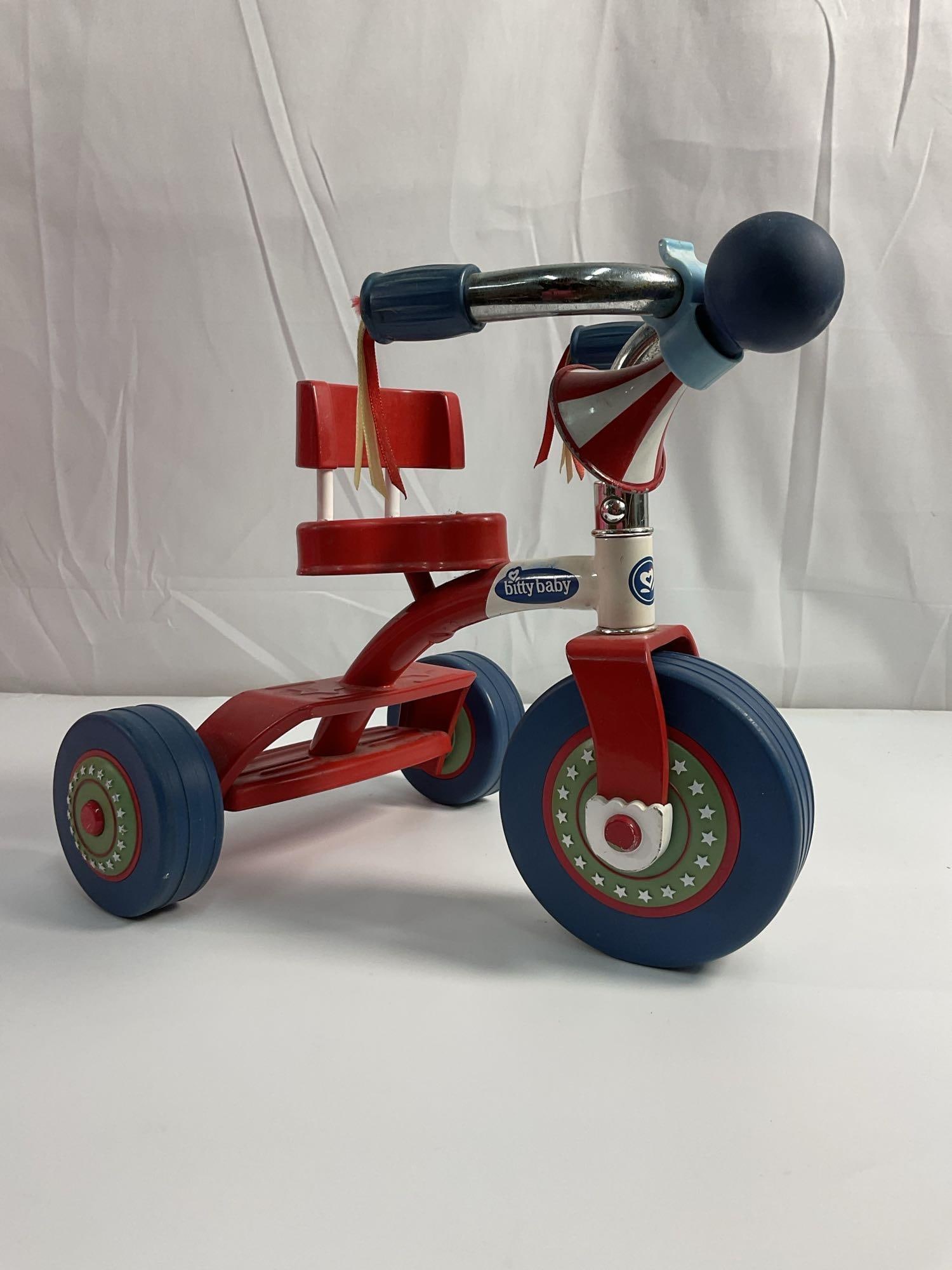 Bitty Baby Tricycle - American Girl: This is a Bitty Baby Tricycle, an accessory designed for Bitty Baby dolls, part of the American Girl doll line. It is a small, red and blue tricycle meant for doll play, featuring