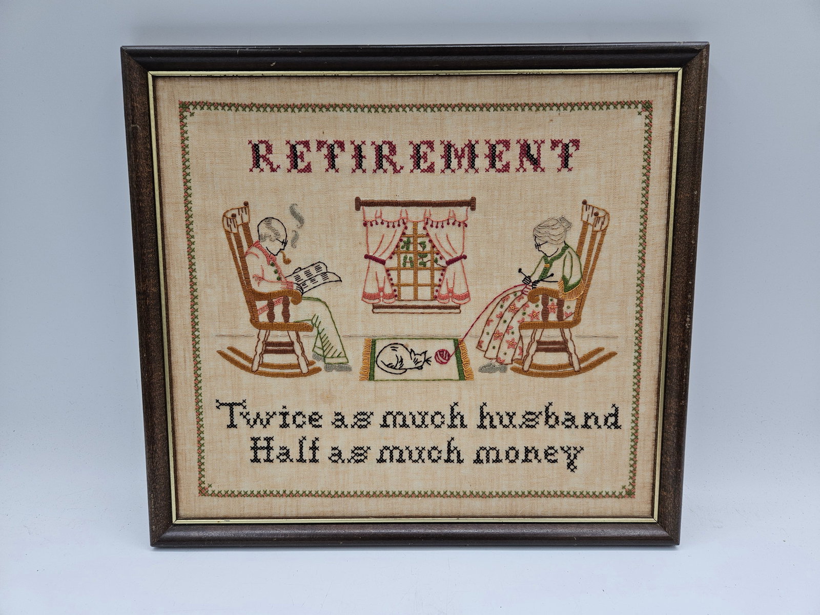 Retirement Sampler Embroidery: Charming framed needlepoint sampler featuring a humorous retirement quote: “Twice as much husband, half as much money.” Depicts a cozy domestic scene with a couple in rocking chairs, embod