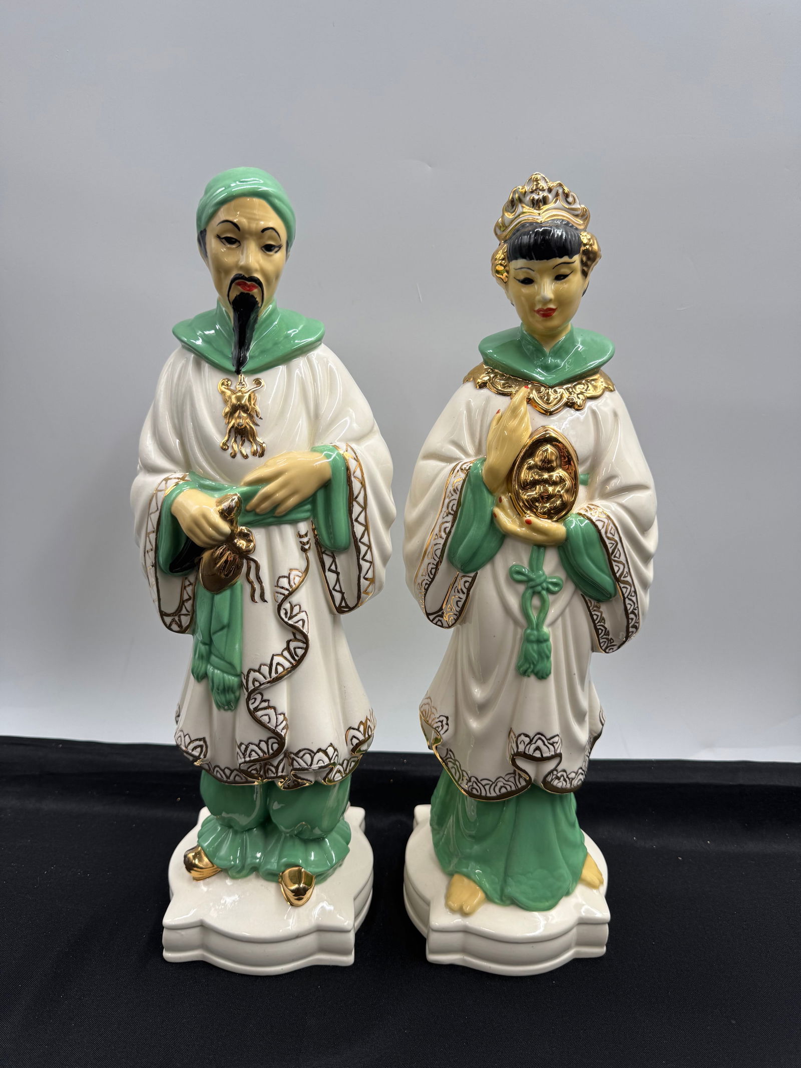 Pair of Vintage Ceramic Asian Figurines: Pair of vintage ceramic figurines depicting Asian figures in traditional attire. The figures are adorned with white robes, green accents, and gold detailing. Each figurine stands approximately 16 inch