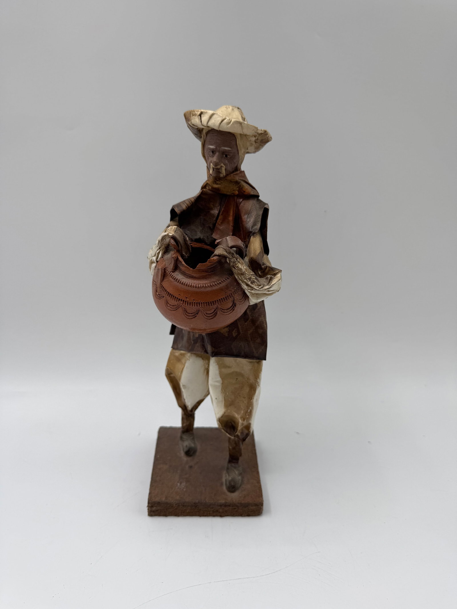 Folk Art Figure of Man with Basket: Folk art figure depicting a man carrying a basket. Constructed of mixed media, possibly paper mache. Mounted on a wooden base. Dimensions: 12.5 x 4 x 4 inches.