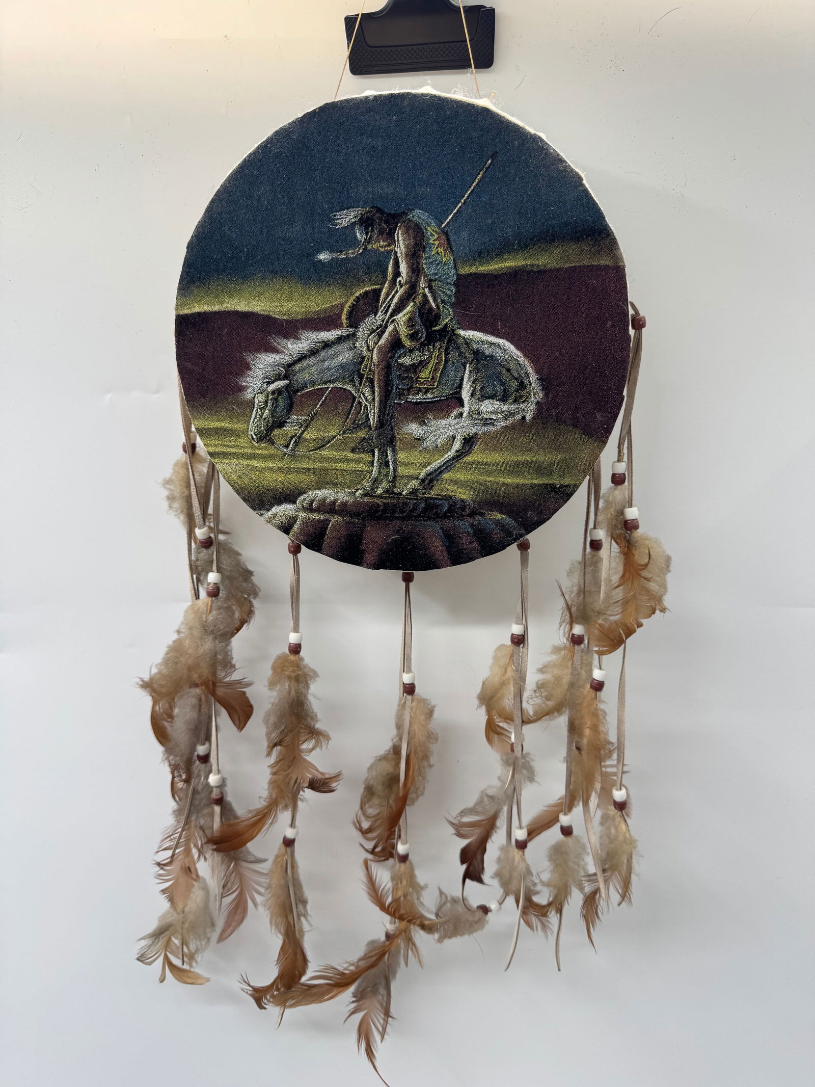 Native American Style Drum with Feathers: Round drum featuring a Native American figure on horseback. Decorated with feathers and beads. Measures approximately 22 x 10 inches.