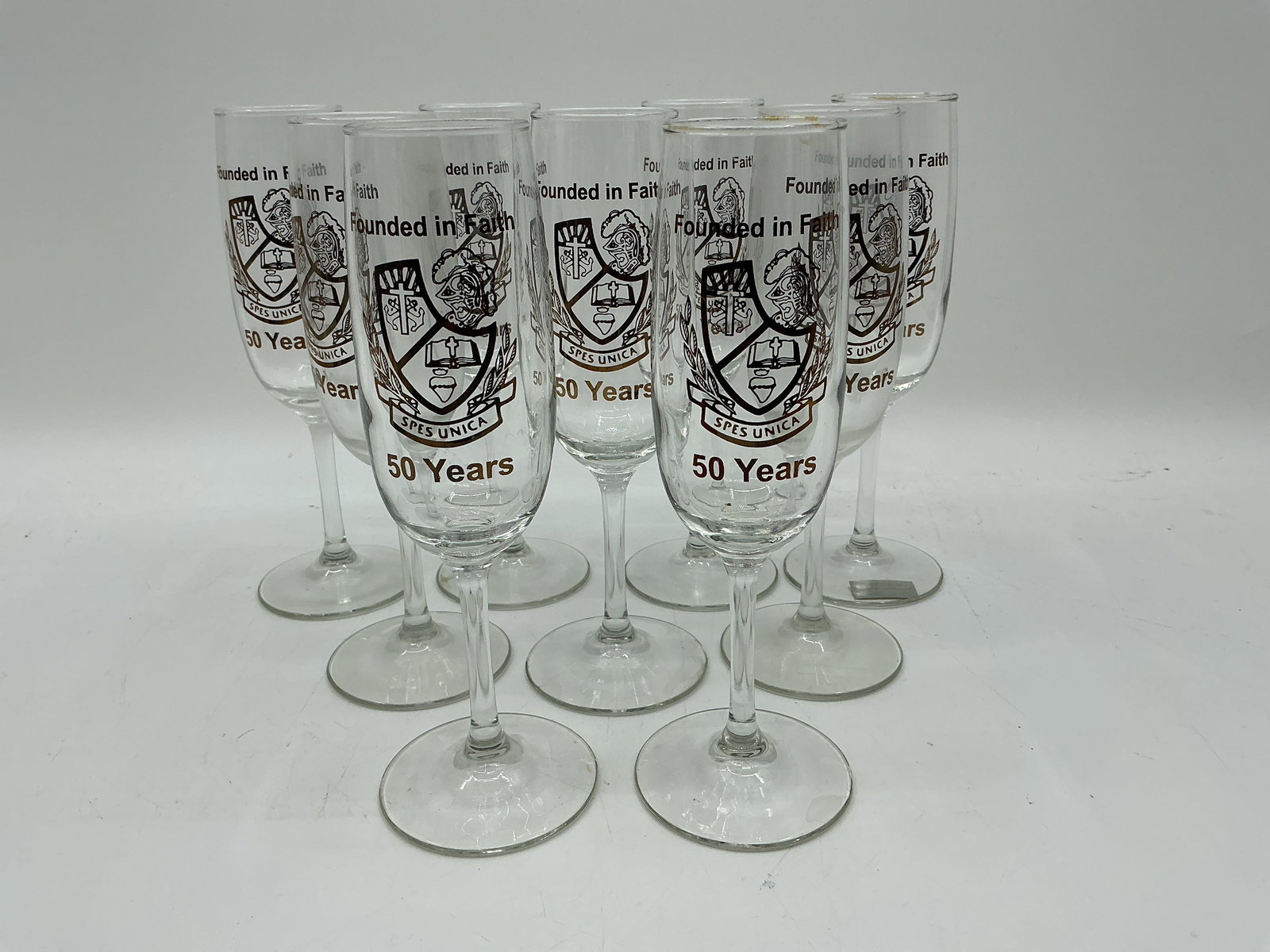 Set of Nine Commemorative Champagne Flutes - SPES Unica: Set of nine champagne flutes, each decorated with a crest and the inscription "Founded in Faith, 50 Years." Each flute measures approximately 8 inches in height.