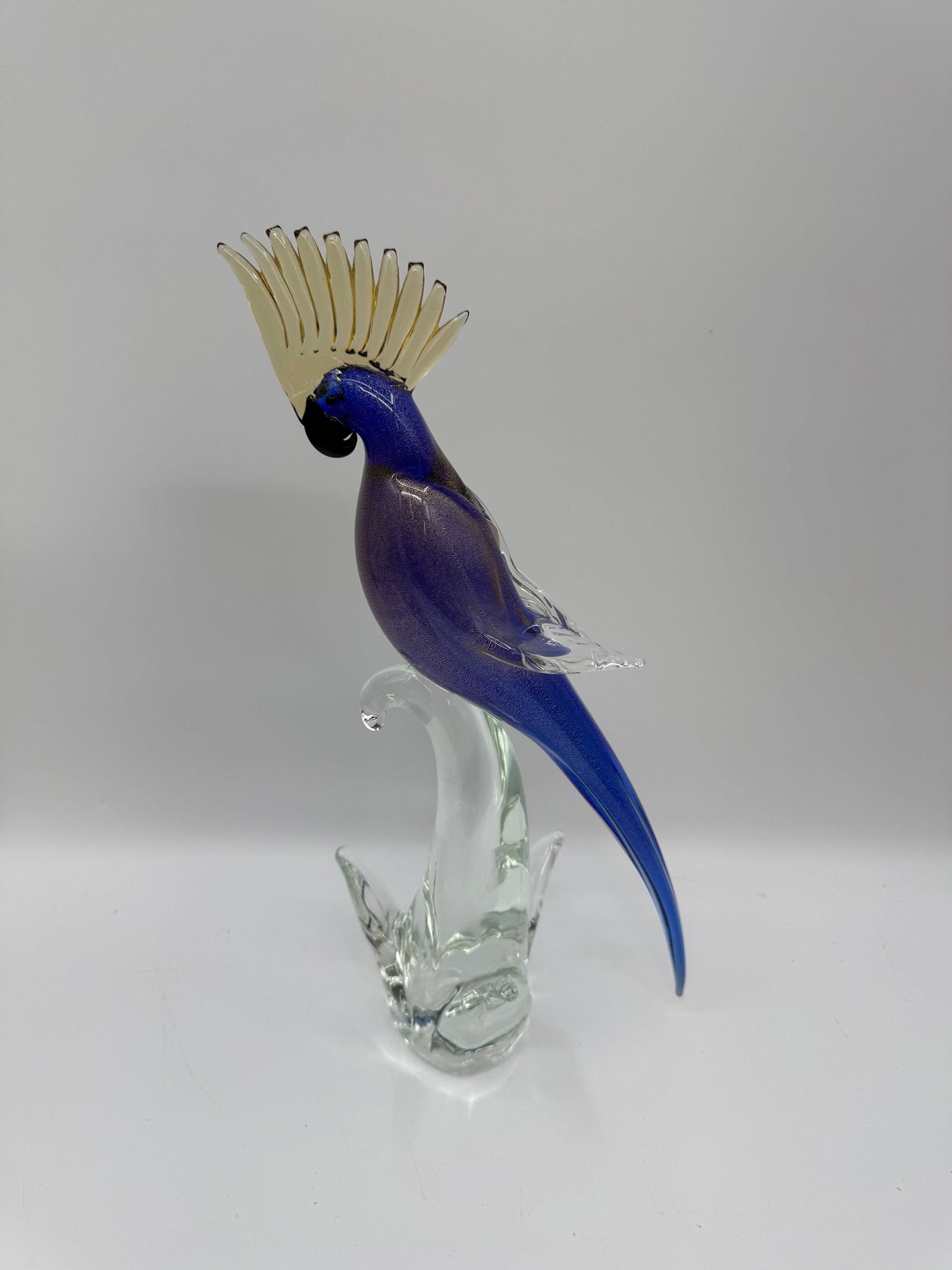 Murano Art Glass Cockatoo Figurine: A striking Murano art glass figurine depicting a cockatoo perched on a clear glass branch. The bird features a vibrant blue body with a distinctive yellow crest. Dimensions: 17 x 9.5 inches.
