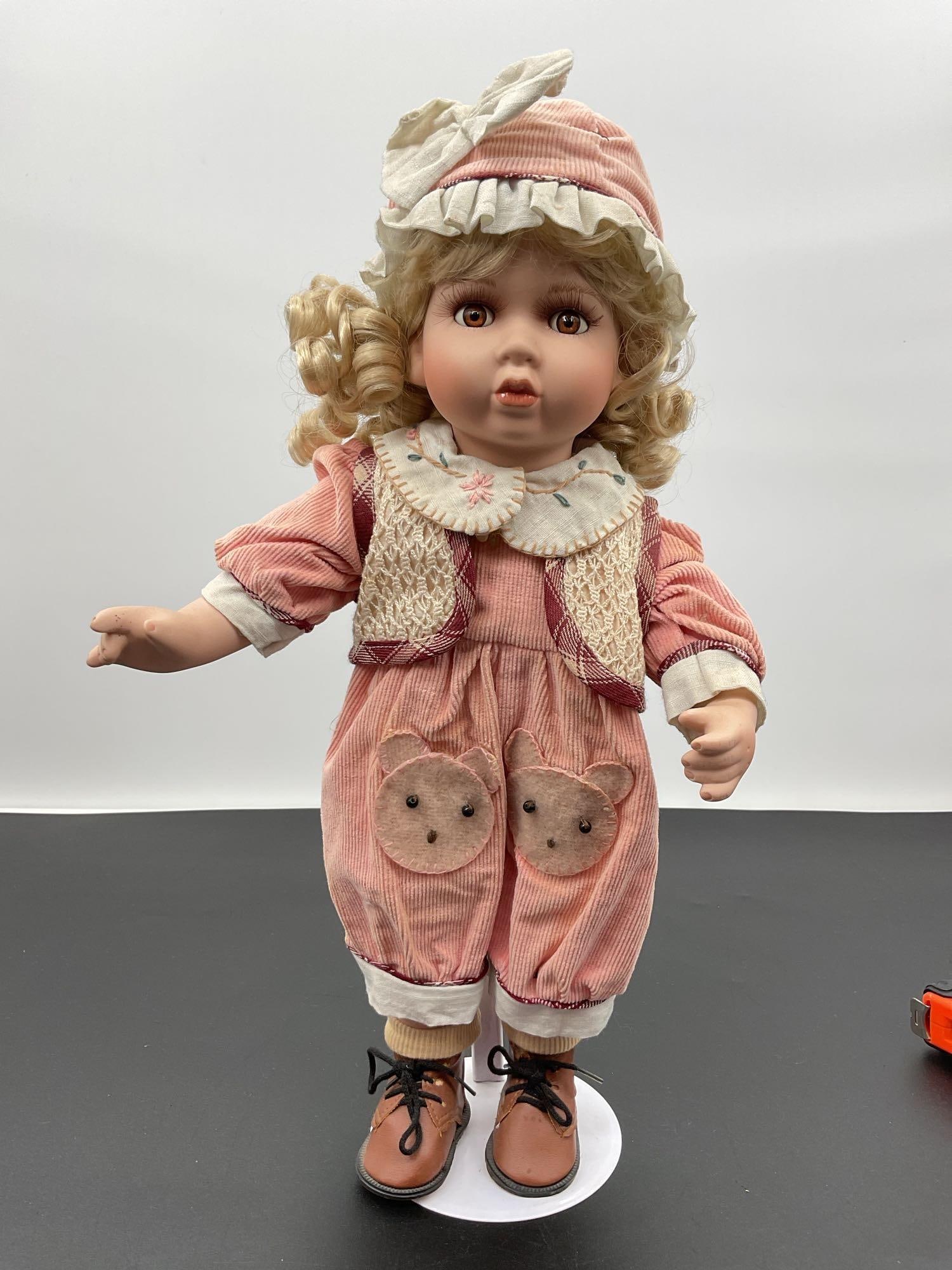 Porcelain Doll in Bear Patch Overalls: This porcelain doll has curly blonde hair, large brown eyes, and a softly surprised expression. She is dressed in a vintage-style outfit featuring pink corduroy overalls with adorable bear face patche