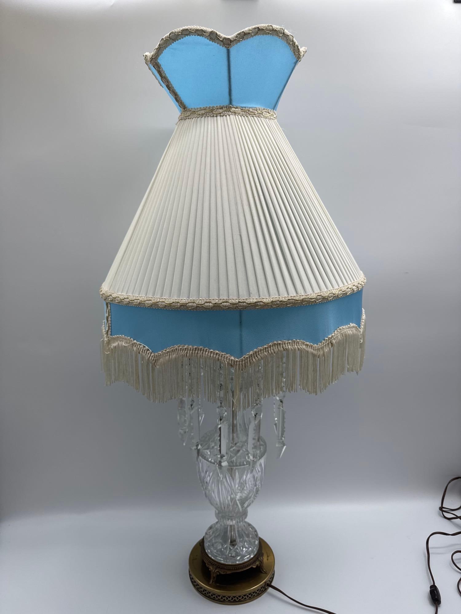 Vintage Victorian Crystal Table Lamp with Prisms and Pleated Shade: Elegant crystal table lamp featuring a faceted cut-glass body with hanging prisms, mounted on a brass base. Completed with a pleated cream shade accented by a blue crown top and fringe trim. Medium Cu