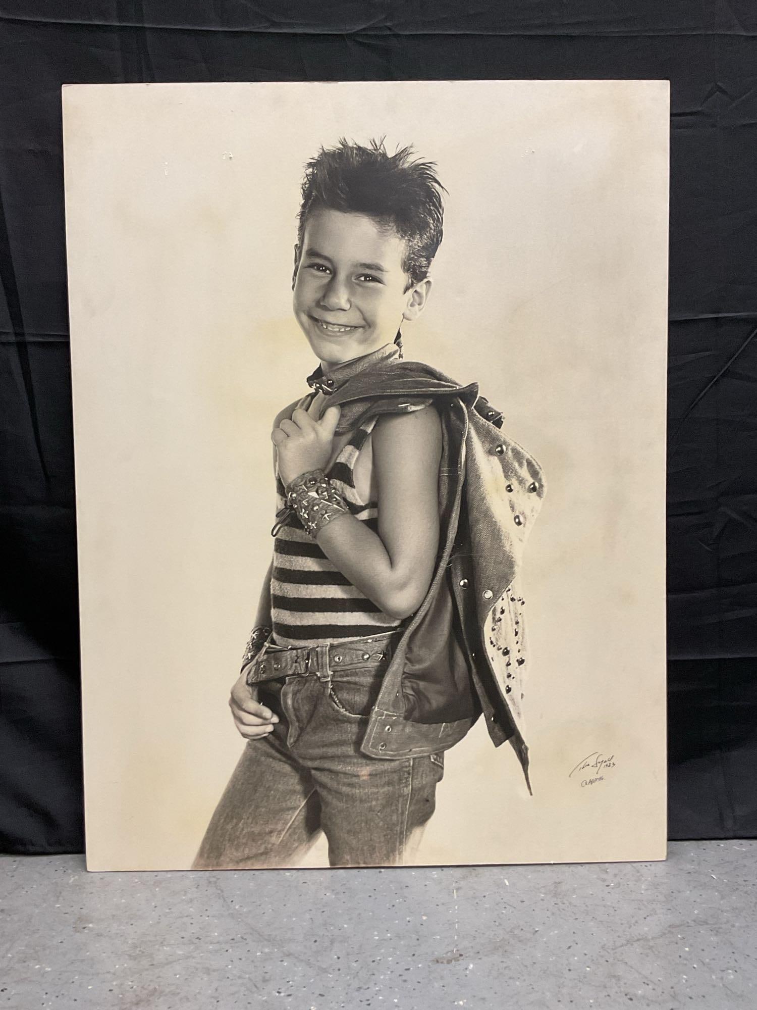 Portrait of a Young Boy by Thea Segall (Romania,1929-2009): Black and white studio portrait by Romanian photographer of a smiling young boy dressed in punk-style fashion, wearing a striped tank top, studded wristbands, and holding a denim jacket ove