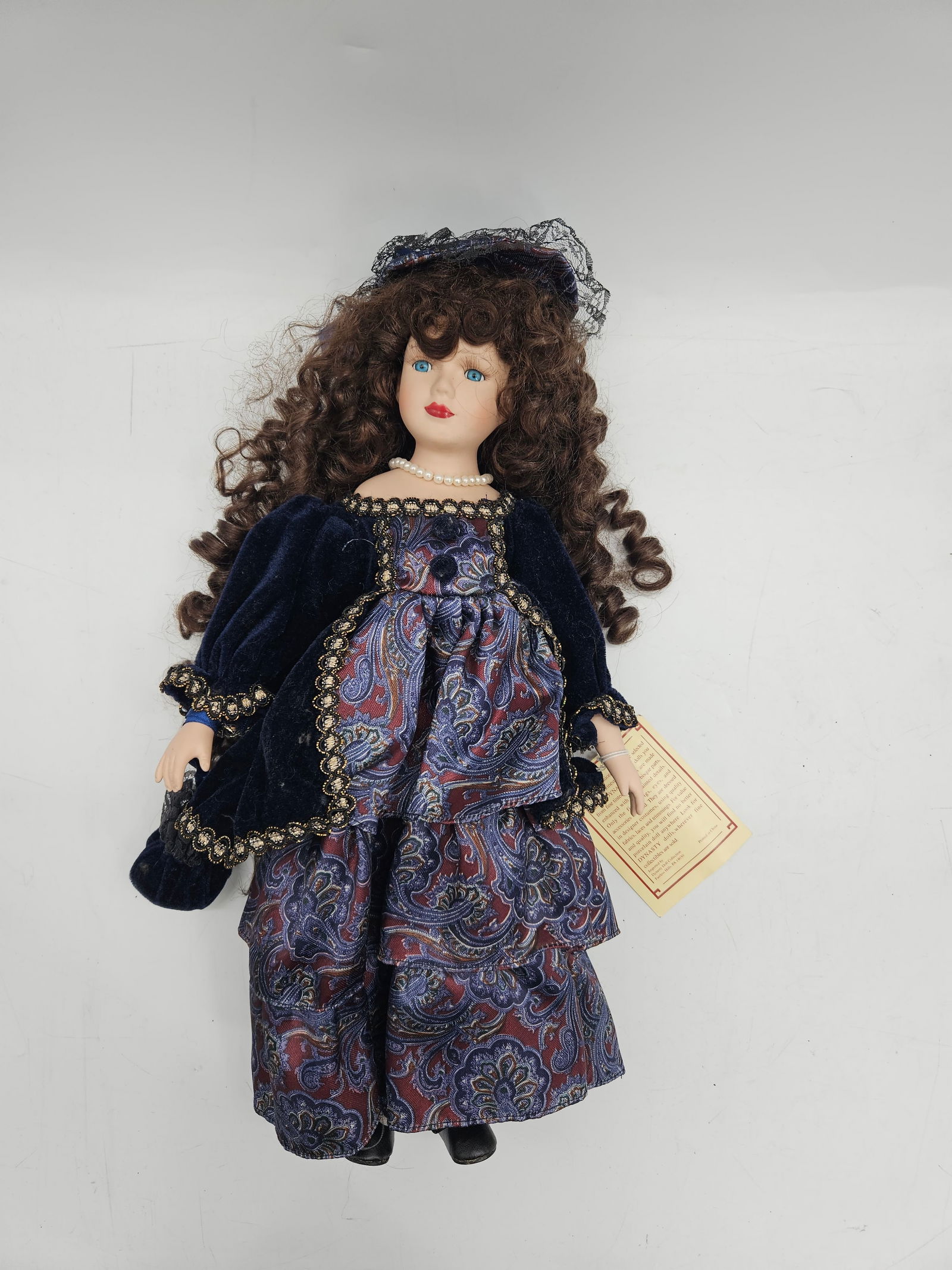 Dynasty Doll Collection Porcelain Doll: Dynasty Doll Collection porcelain doll with hand-painted details, designer costume, and curly brown hair. Includes original tag. Measures approximately 17 inches tall and 9 inches wide.