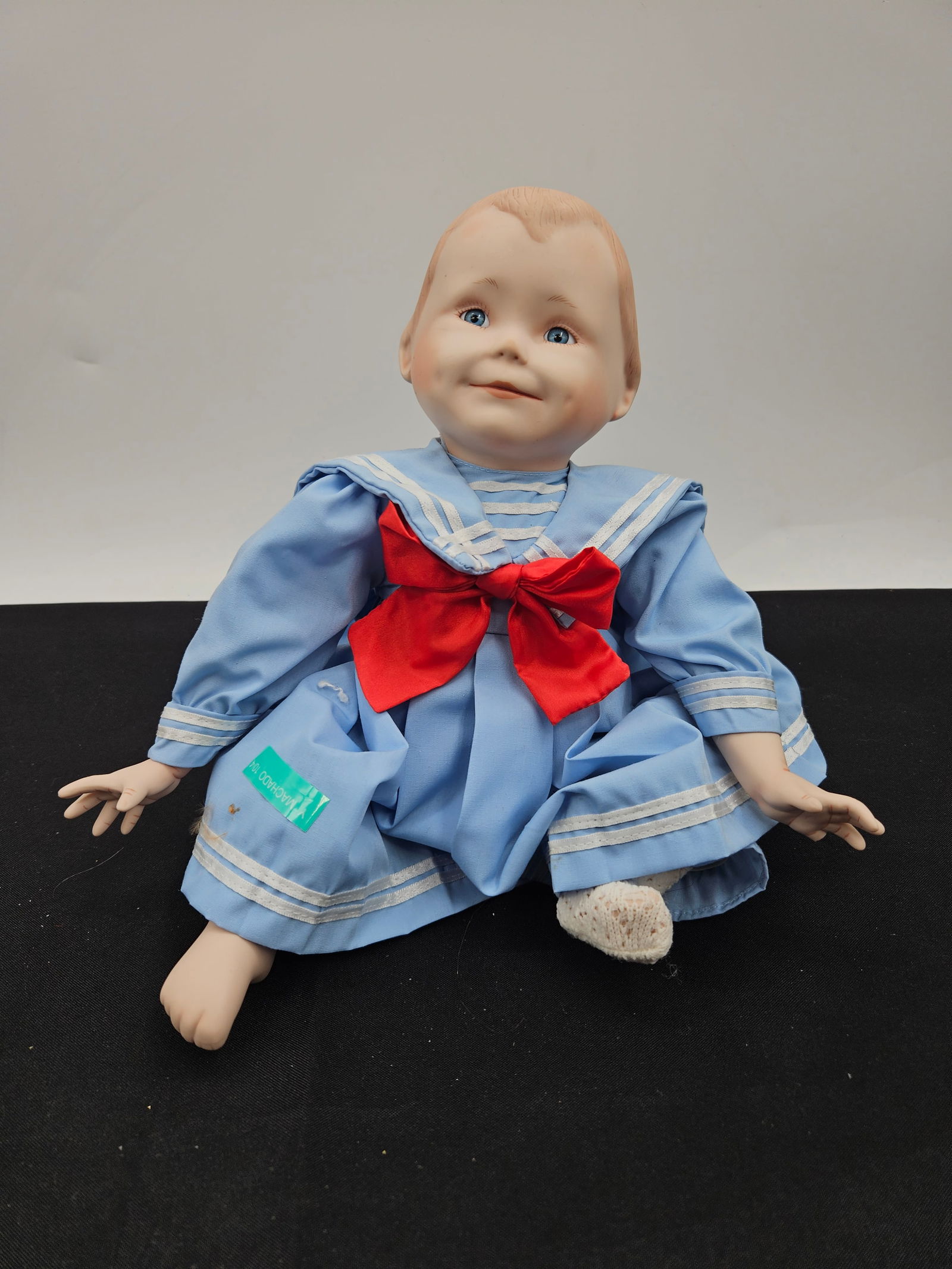 Porcelain Doll in Sailor Suit: Porcelain doll with painted features, dressed in a blue sailor suit with a red bow. Measures approximately 9 inches high, 10 inches wide at the widest part, and 5 inches deep.