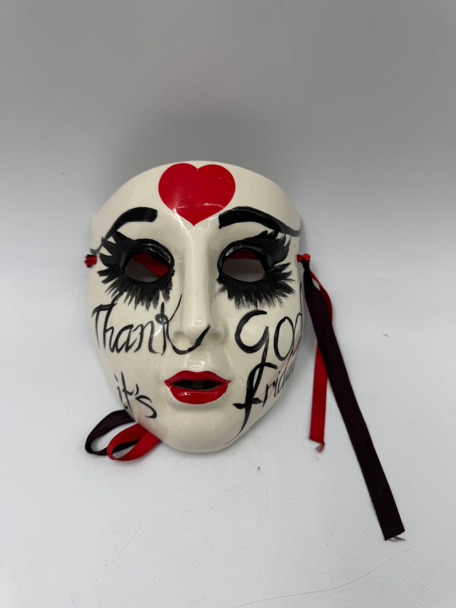 Hand-Painted Ceramic Mask, "Thank God It's Friday": Hand-painted ceramic mask featuring a stylized face with a red heart on the forehead and the inscription "Thank God It's Friday". The mask has black eyelashes and red lips, with ribbon