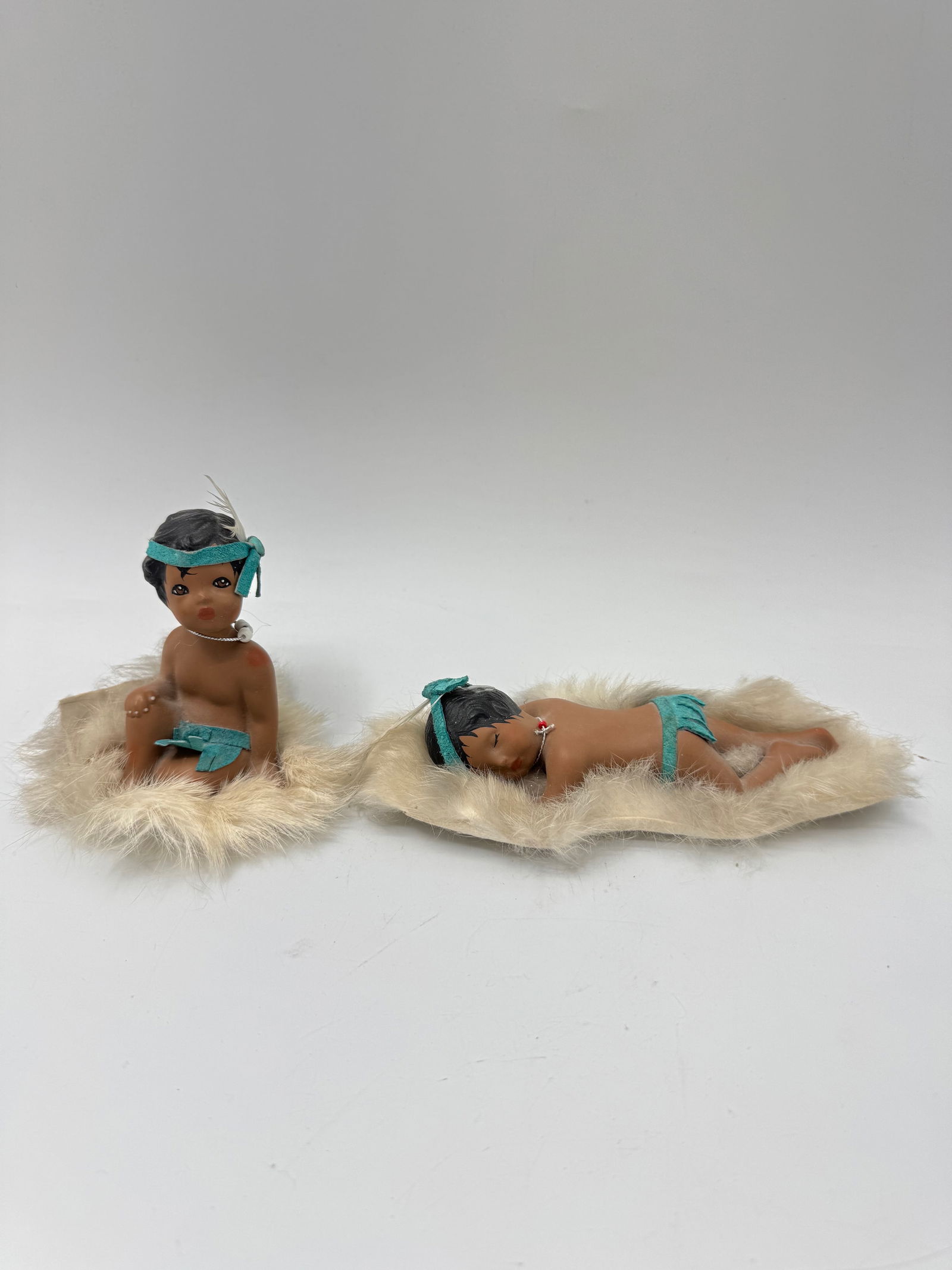 Two LTM 55 Evening Star Native American Dolls: Two LTM 55 Evening Star Native American dolls. One is seated, and the other is lying down. Each doll is approximately 6 inches in length and is displayed on a faux fur base.