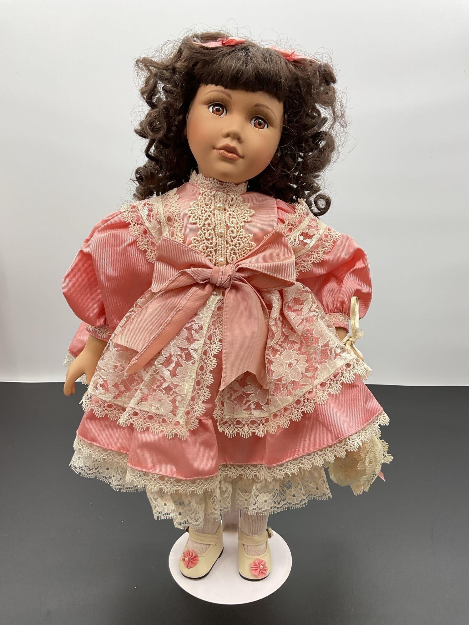 Porcelain Doll in Pink Satin and Lace Dress: This elegant porcelain doll features dark curly hair and a gentle, expressive face. She is dressed in a soft pink satin dress accented with detailed ivory lace overlay and a large matching bow at the