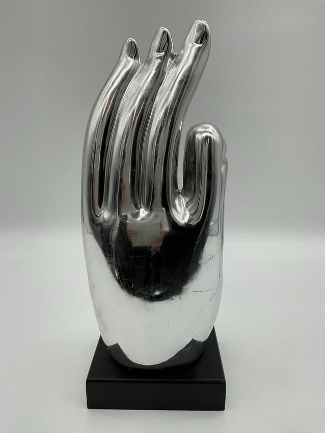 Silver-Tone Ganesha in Hand Sculpture: Contemporary decorative sculpture featuring the Hindu deity Ganesha seated within an open hand, symbolizing protection and blessings. The piece has a polished silver-tone finish and rests on a black r