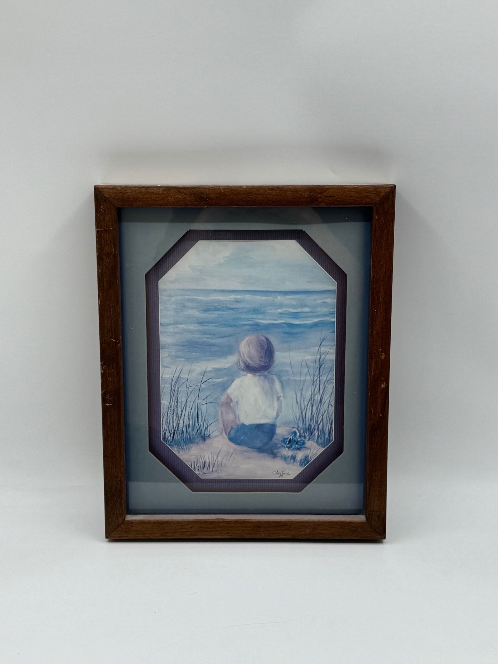 Clifford, Boy at the Beach, Framed Print: Framed print depicting a young boy sitting on a beach, gazing out at the ocean. Signed "Clifford" in the lower left. Image size approximately 15 x 12 inches. Framed and ready to hang.