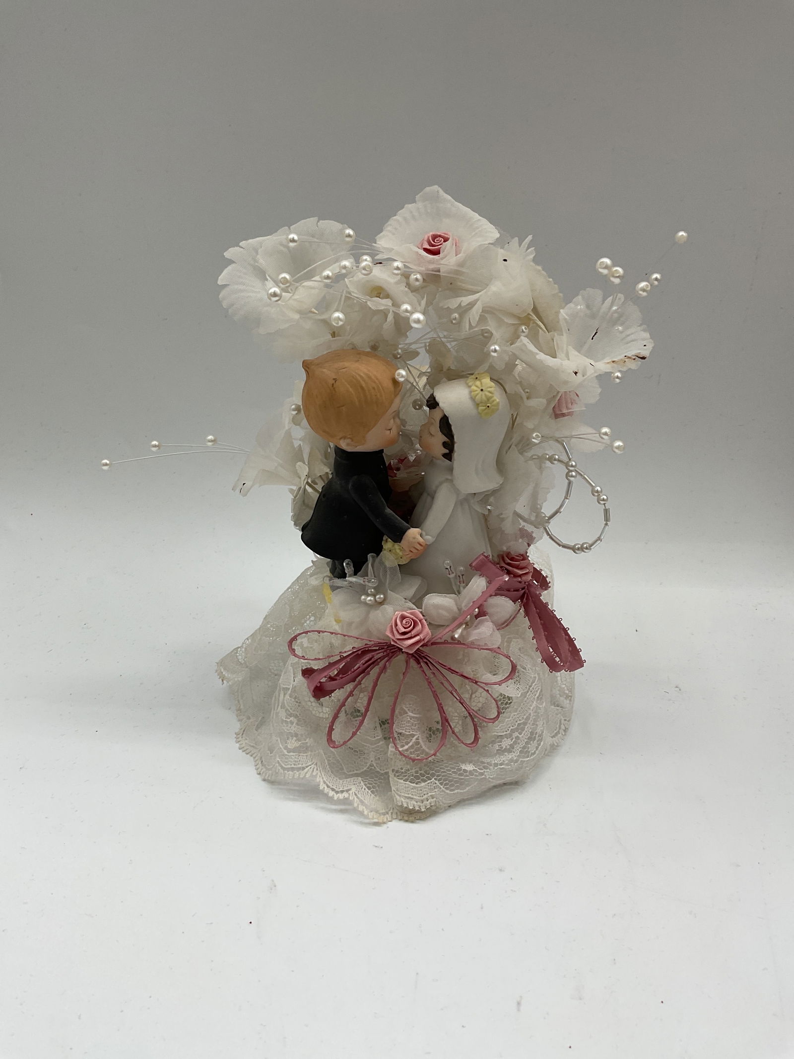 Vintage Light-Up Bride and Groom Cake Topper: Vintage wedding cake topper featuring a bride and groom embracing beneath lace, ribbon, and pearl decorations. The piece includes a built-in battery compartment that illuminates the floral