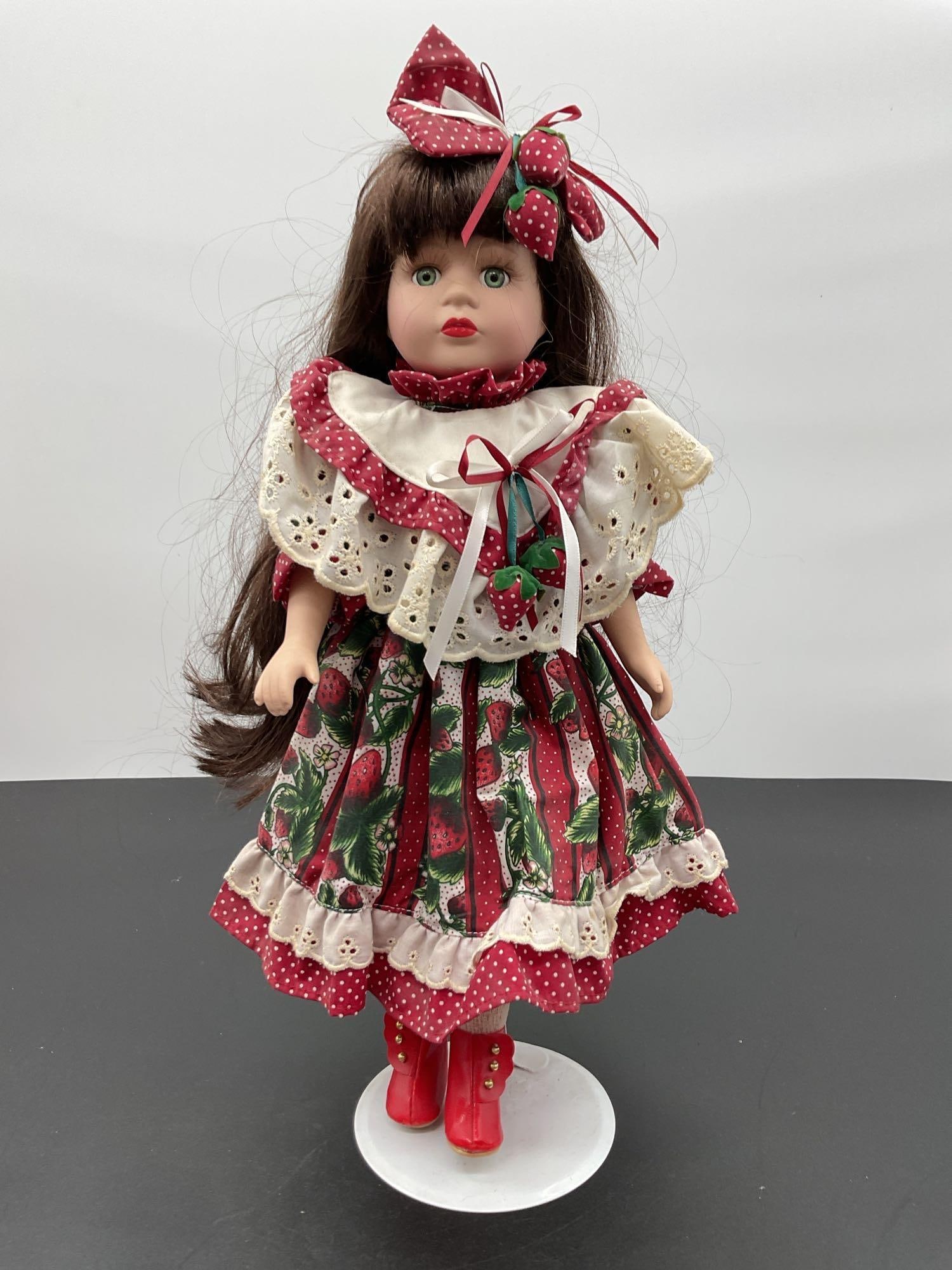 Porcelain Doll in Strawberry Print Dress with Red Boots: This vibrant porcelain doll features long dark brown hair, striking green eyes, and bright red lips. She is dressed in a red and green strawberry-themed dress with a white eyelet lace collar, matching