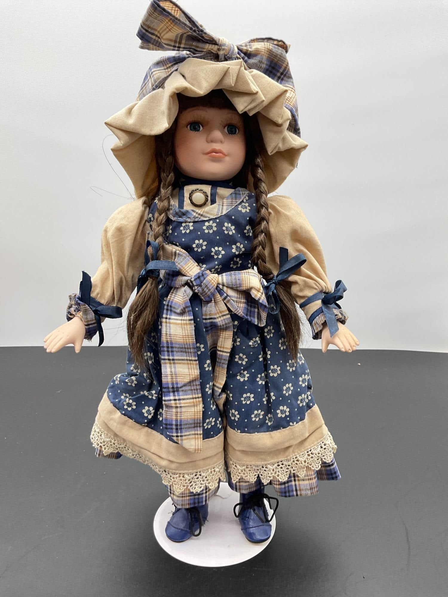 Porcelain Doll in Blue Prairie Dress with Bonnet: This porcelain doll features long brown braids, soft blue eyes, and a calm expression. She wears a prairie-style dress in navy blue with small white floral prints, beige accents, and a plaid apron