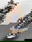 Porcelain Doll in Blue Prairie Dress with Bonnet: This porcelain doll features long brown braids, soft blue eyes, and a calm expression. She wears a prairie-style dress in navy blue with small white floral prints, beige accents, and a plaid apron