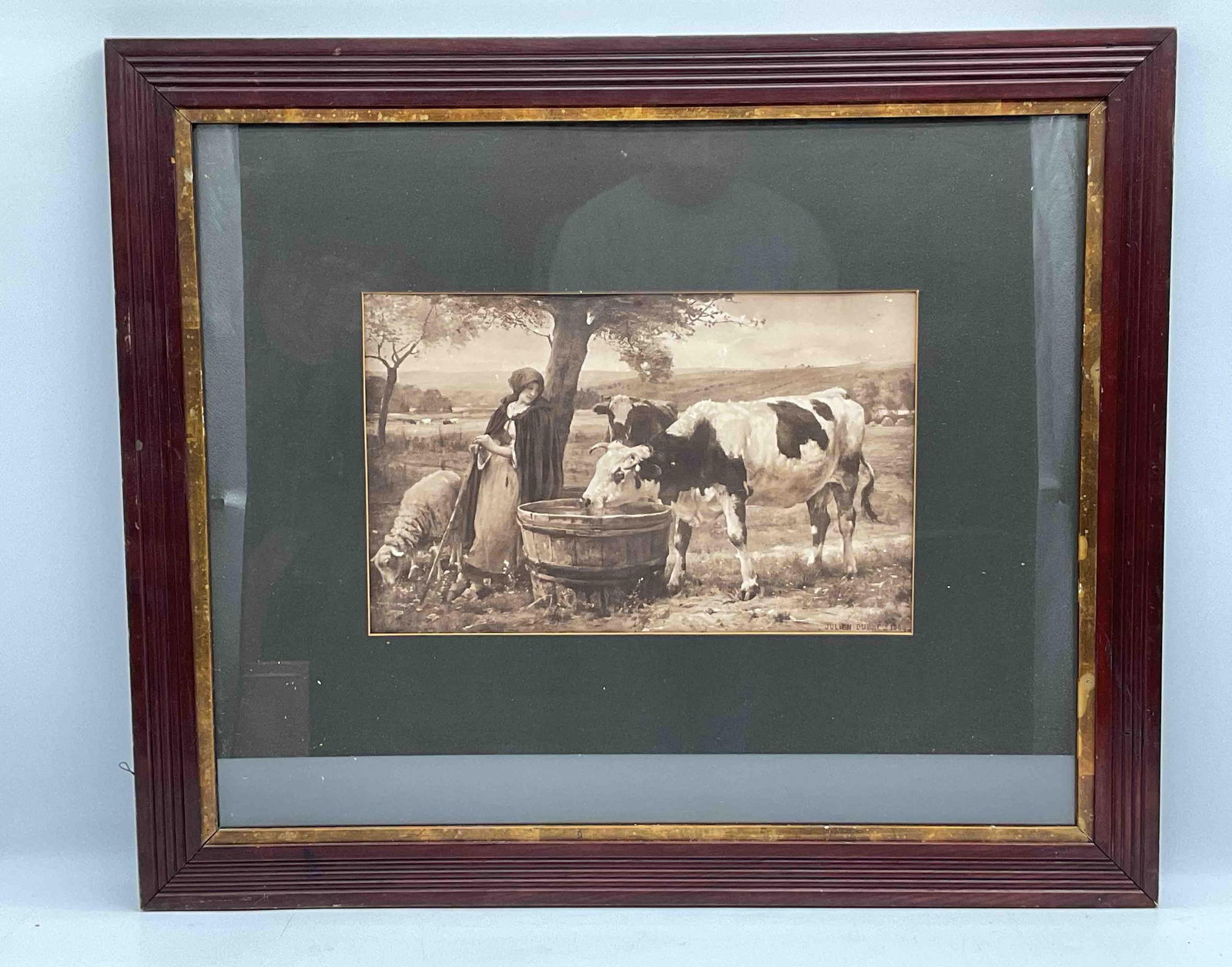 Antique Sepia Print of Farm Scene with Cows and Shepherdess: Early 20th-century sepia-toned print depicting a pastoral farm scene with a young shepherdess, a cow drinking from a trough, and a sheep nearby. Framed under glass in a dark wood frame with gilt