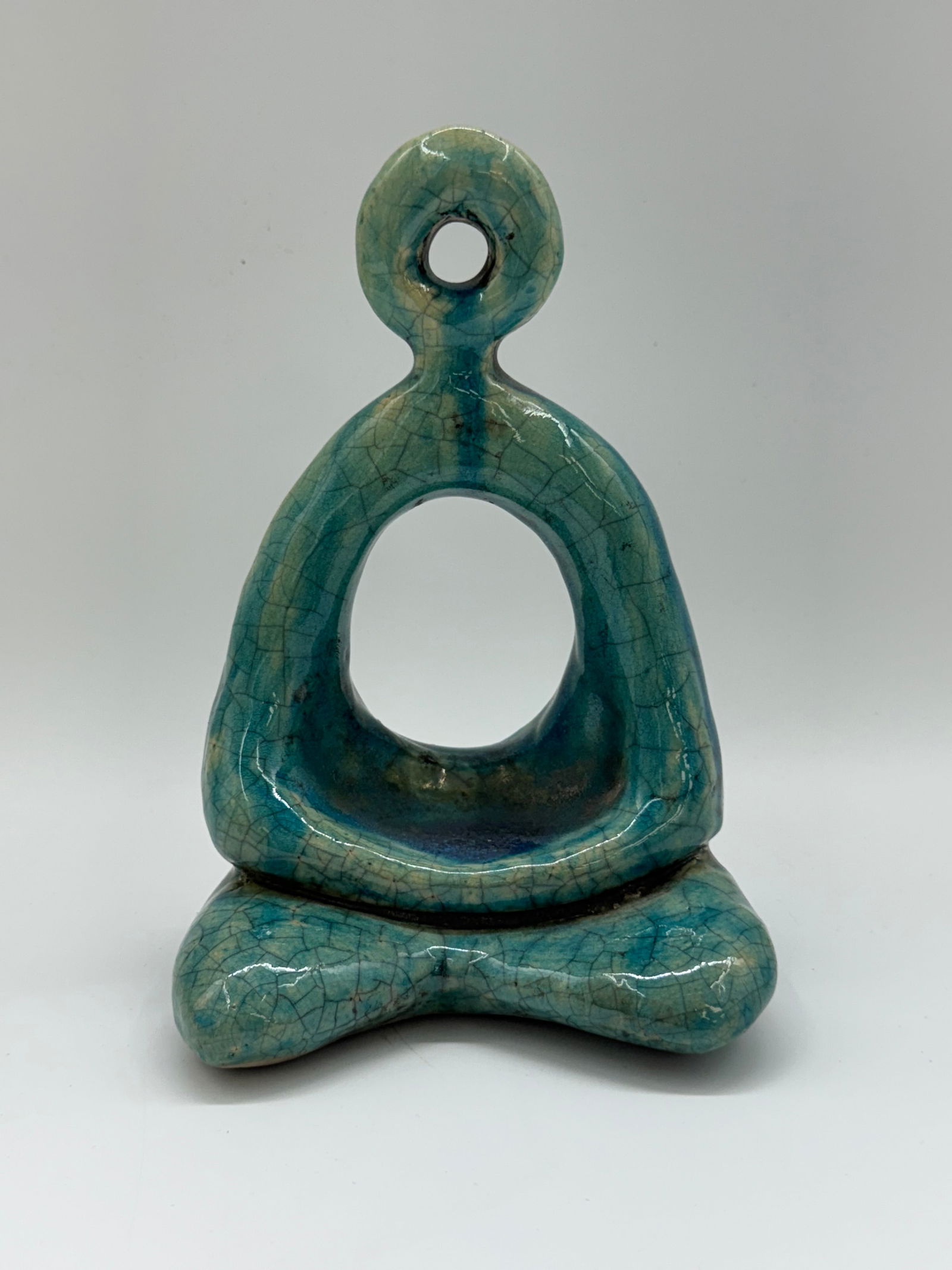 Modern Turquoise Ceramic Abstract Meditation Figure: Stylized modern ceramic sculpture depicting an abstract seated figure in a meditative pose. The piece features a smooth, flowing form with an open circular center and a looped head, emphasizing harmon