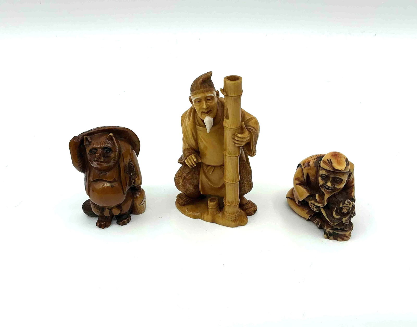 Three Japanese Style Netsuke Figurines: Carved figures including a tall standing elder with staff a crouching figure with a mask and a seated figure holding an object likely crafted from resin or bone imitation material Medium Carved resin