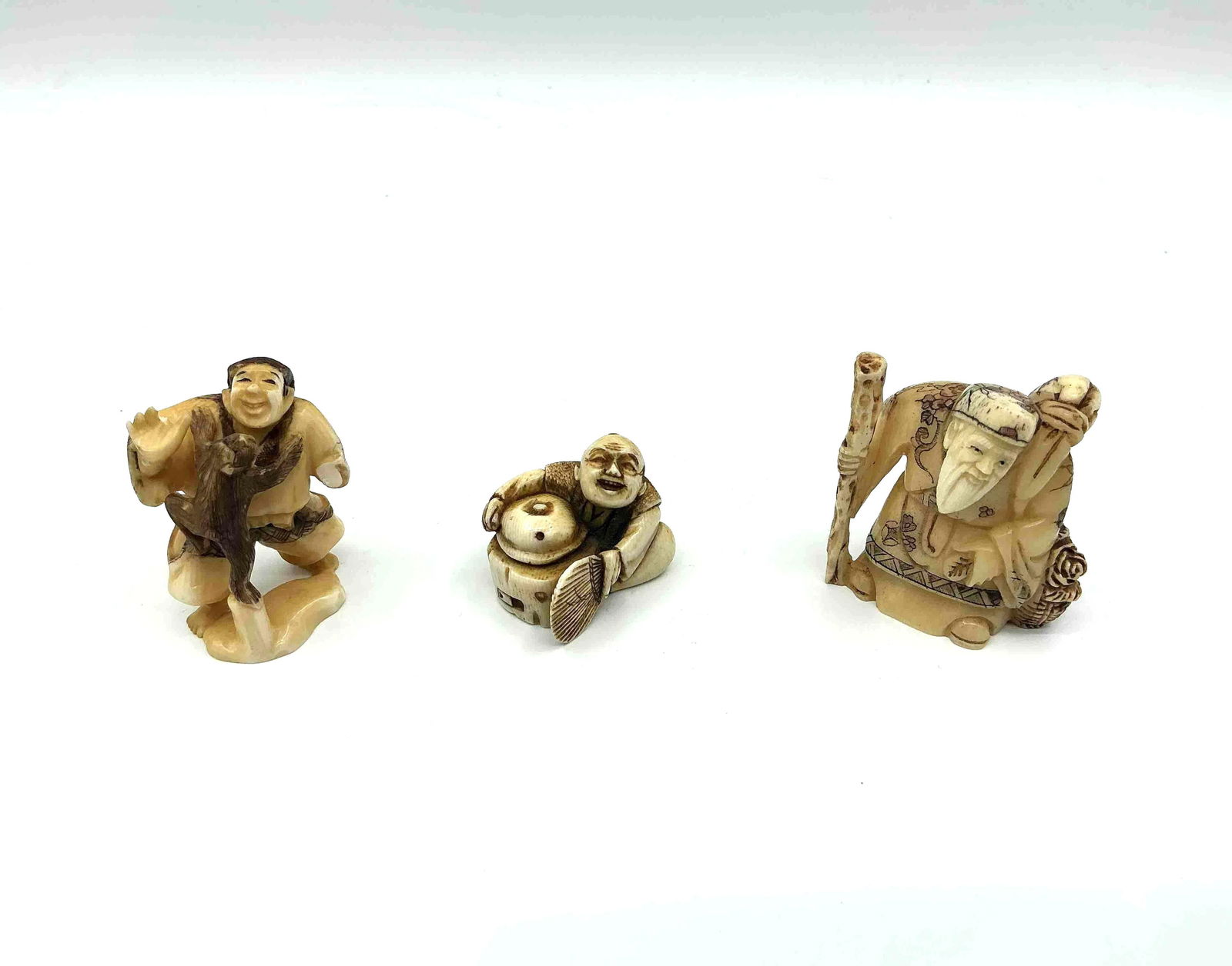 Three Japanese Style Netsuke Figurines: Trio of carved figures including an elder with staff a man carrying a sack and a seated character holding a mask likely crafted from resin or bone imitation material Medium Carved resin or composite