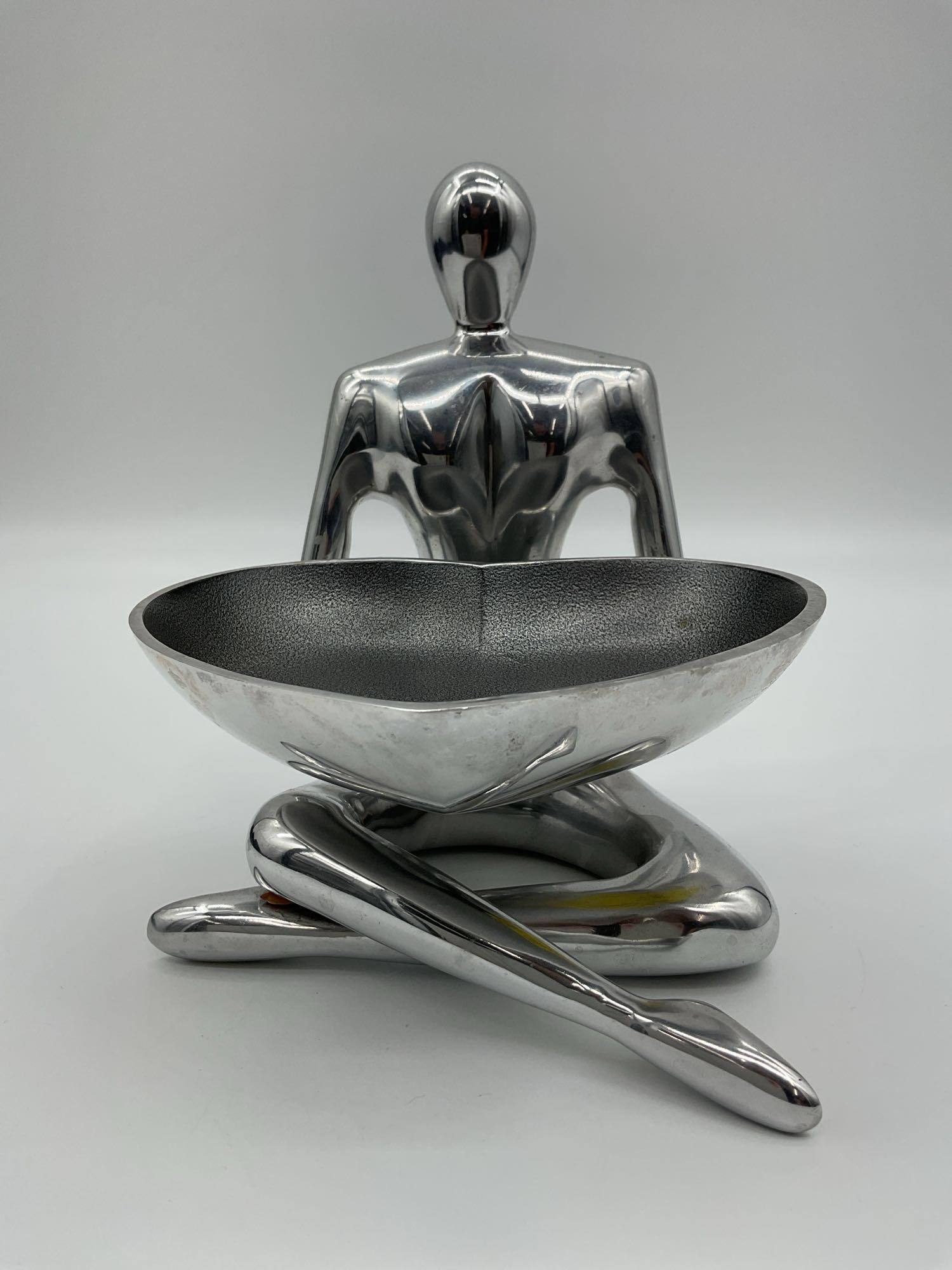 Nambe Heart Bowl with Abstract Metal Figure Stand: Modernist style heart-shaped bowl by Nambe, held by a sleek abstract aluminum figure in a seated pose. The piece blends functional design with sculptural art, ideal for serving or decorative