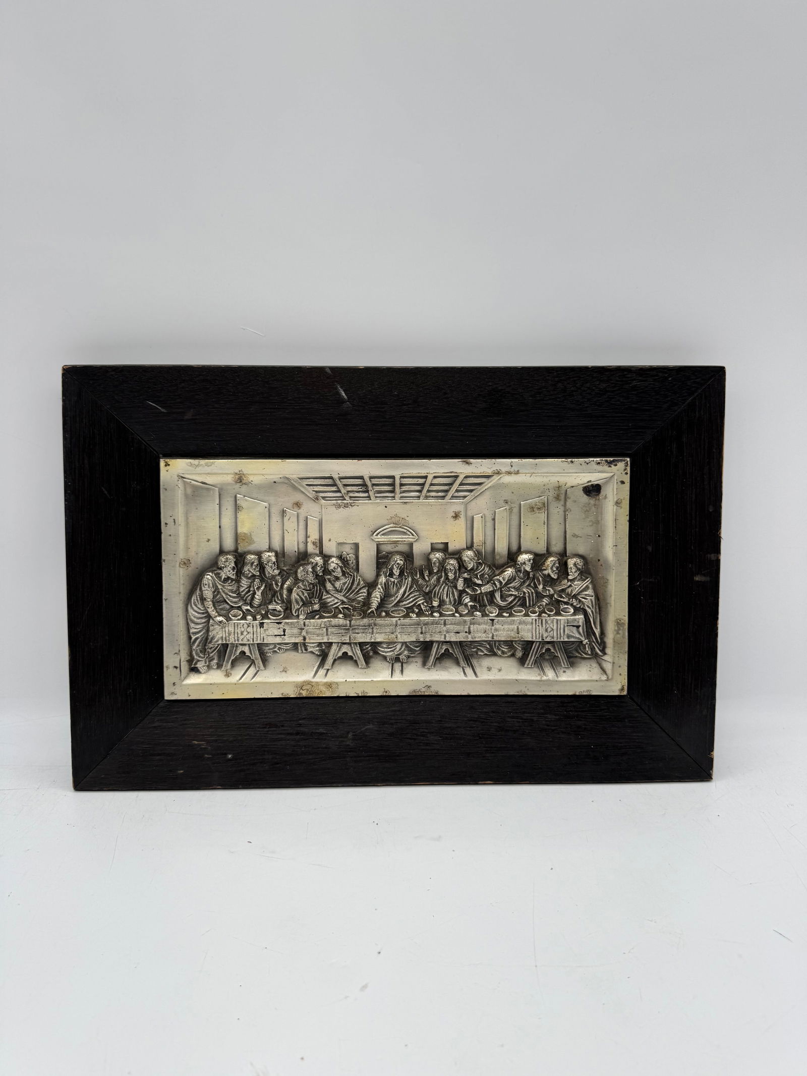 The Last Supper Relief Plaque: Framed relief plaque depicting The Last Supper. The plaque is metal and set within a dark wooden frame. Dimensions: H:11in W:18in 