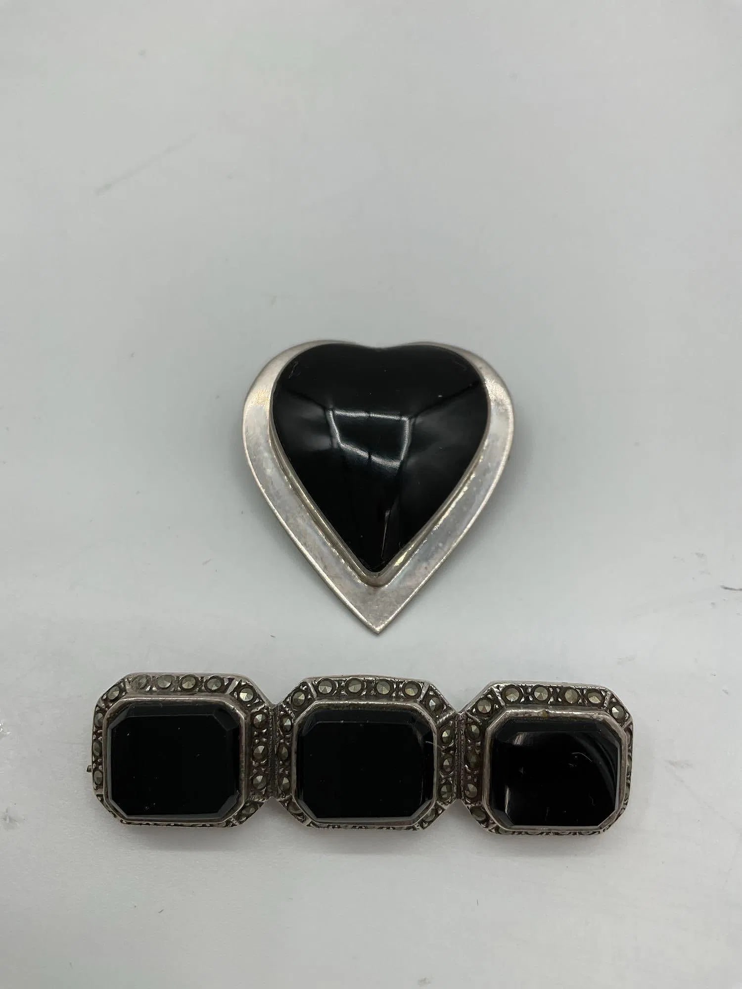 Pair of Vintage Onyx and Sterling Silver Brooches: Includes one heart-shaped brooch with polished black onyx set in a silver tone frame, and one rectangular triple-stone brooch with faceted black stones framed by marcasite-style detailing. stamped 925