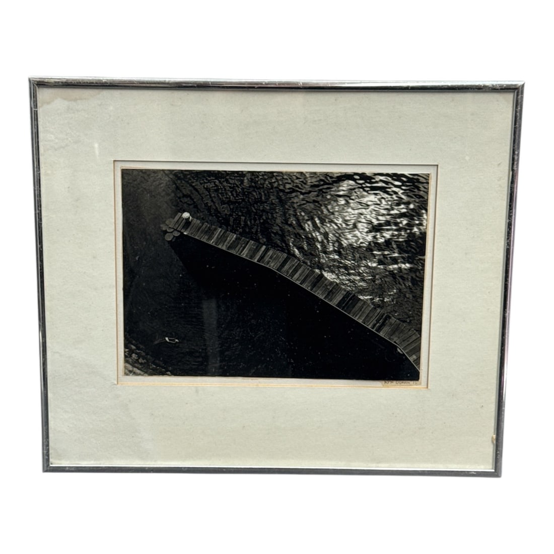 Framed Black and White Photograph by Ron Olman, 1975: This black and white photograph by Ron Olman, dated 1975, presents a striking overhead view of a narrow wooden walkway extending across dark, reflective water. The composition emphasizes strong diagon