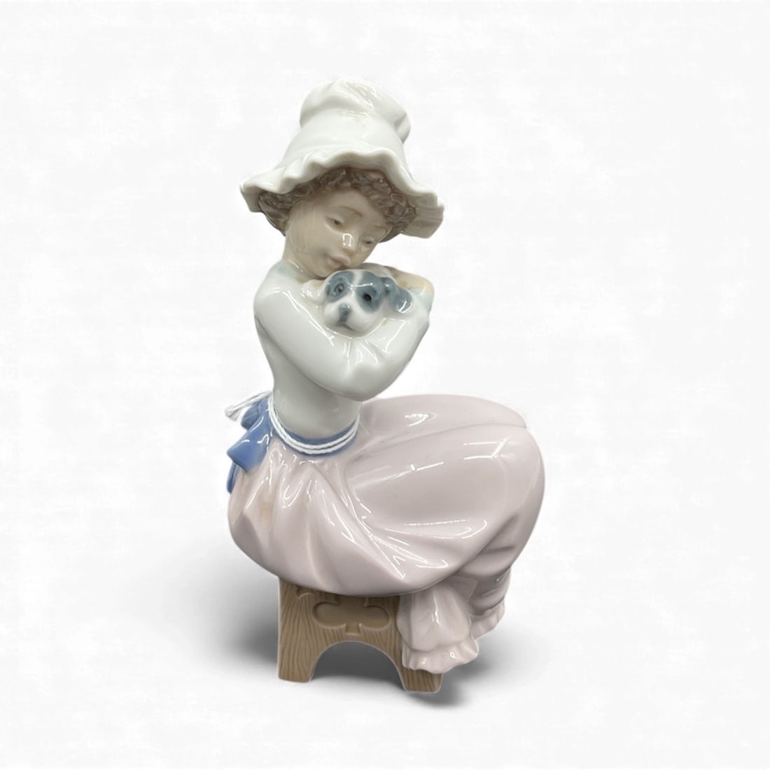 Nao by Lladro Girl with Puppy Figurine: A charming Nao by Lladro porcelain figurine featuring a young girl in a wide bonnet seated on a small stool while gently holding a puppy in her arms. Rendered in soft pastel tones with a glossy glaze