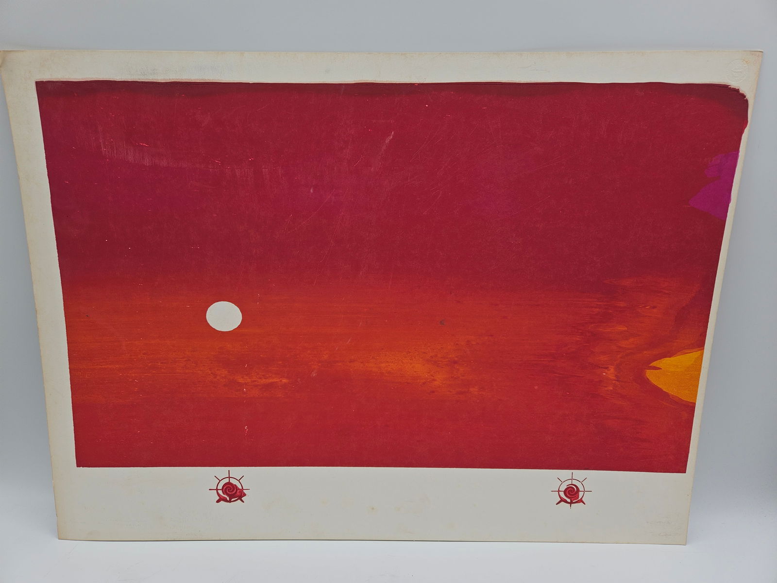 Abstract Red Landscape Print: Abstract landscape print featuring a red and orange sky with a white circle, possibly a sun or moon. The print is mounted on a white backing with decorative red emblems along the bottom. Dimensions: 2
