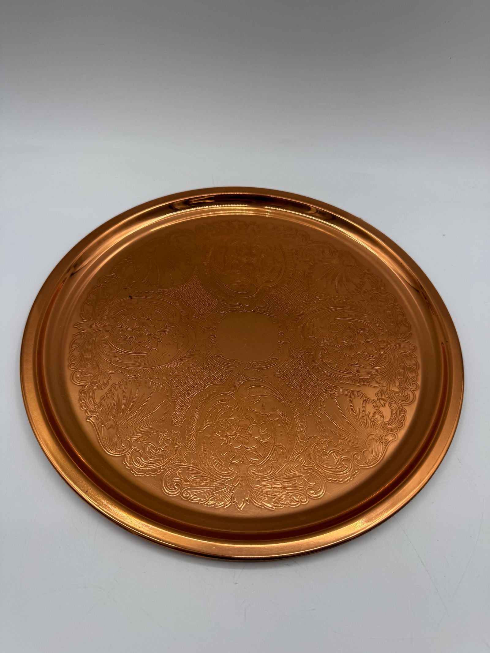 Engraved Copper Tray, 12 inch Diameter: A circular copper tray featuring engraved floral and paisley designs. The tray measures approximately 12 inches in diameter and has a raised rim. The reverse is plain copper.