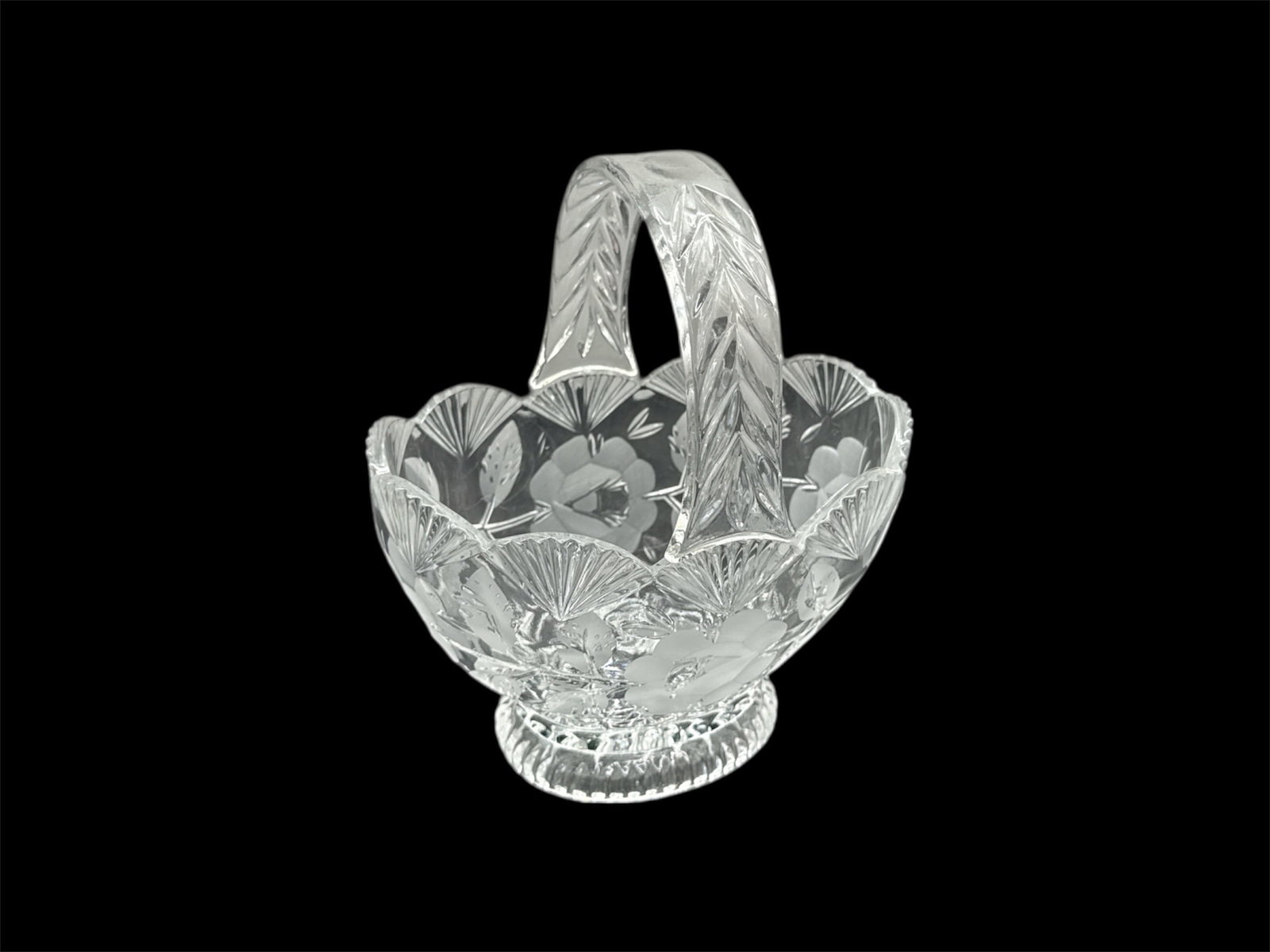 Cut Glass Handled Basket with Floral Etching: Clear cut glass basket featuring a scalloped rim and integrated handle, decorated throughout with etched floral and foliate motifs. The bowl rests on a faceted pedestal base, adding visual height and