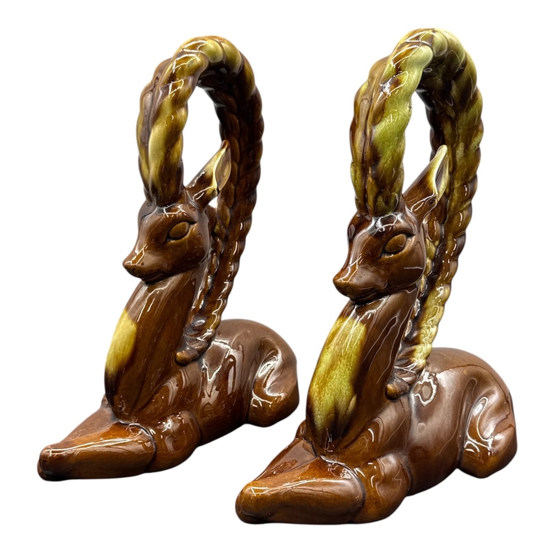 Pair of 1950s Ceramic Ram Sculptures: An uncommon pair of ceramic ram sculptures dating to the 1950s, each modeled in a reclining pose with elongated necks and dramatic braided horn forms arching overhead. Finished in rich brown and green