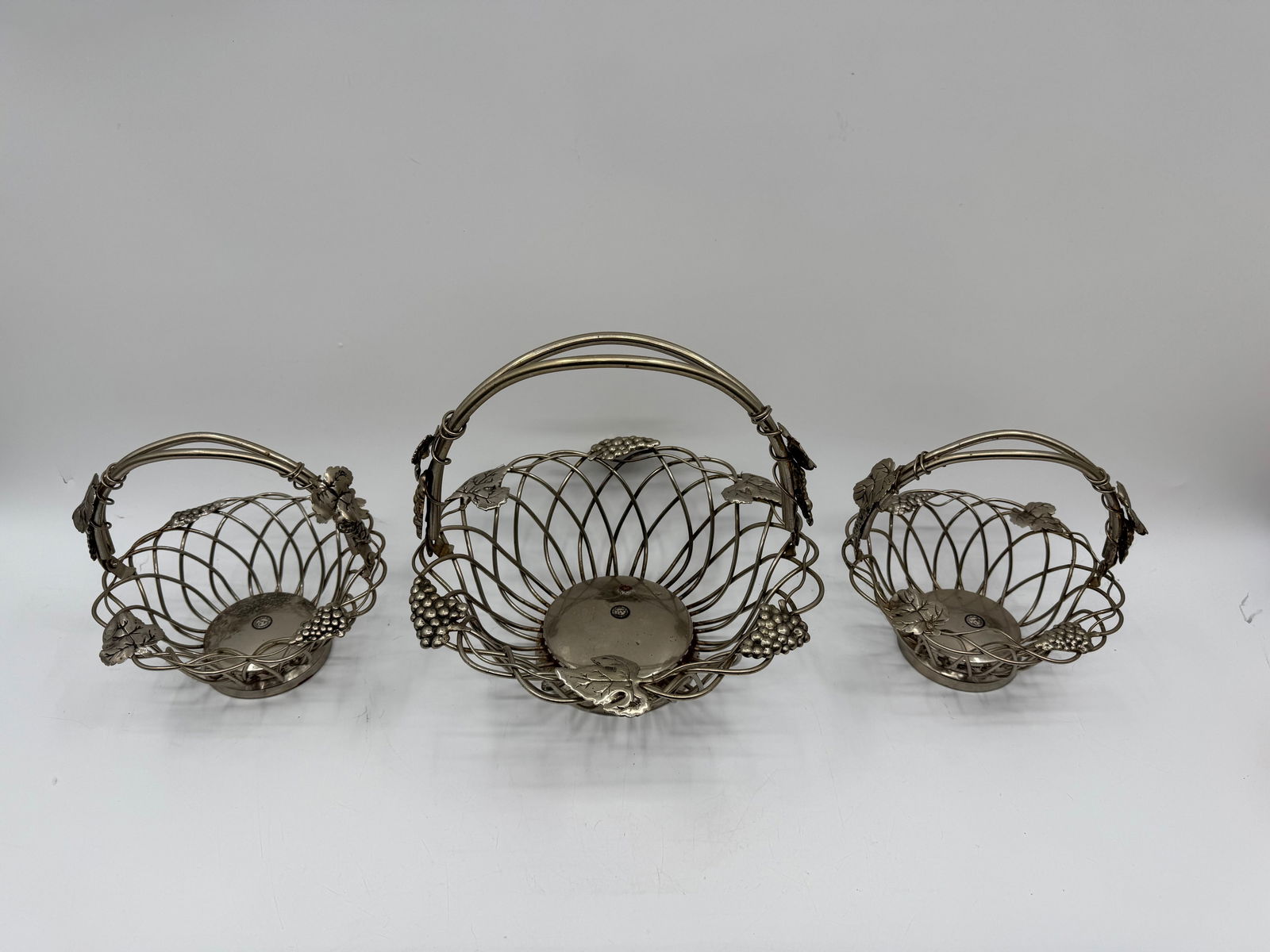 Set of Three Godinger Silver Art Co. Baskets: Set of three silver-tone metal baskets by Godinger Silver Art Co. Features decorative grape and leaf accents on the handles. Sizes vary, with the largest approximately 11 inch D 10 inch H.
