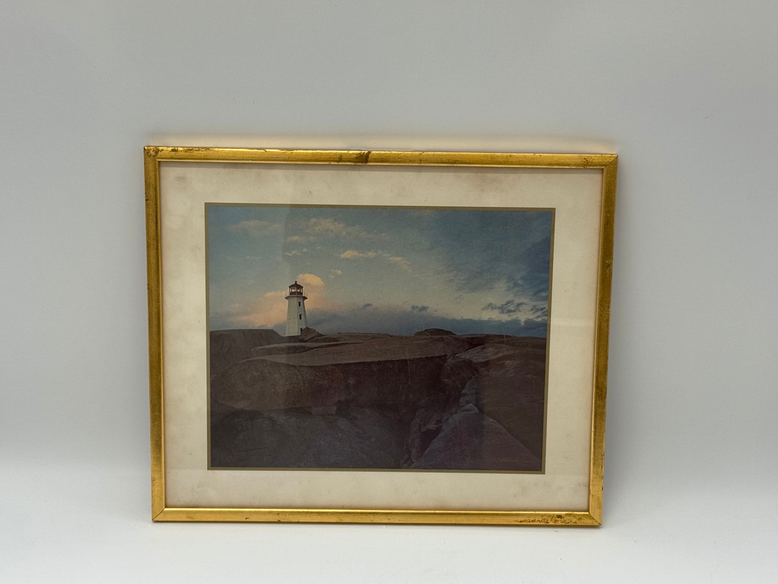 Lighthouse at Sunset, Framed Print: Framed print depicting a lighthouse on a cliffside at sunset. Matted and presented in a gold-tone frame. Dimensions: 17 x 14 inches.