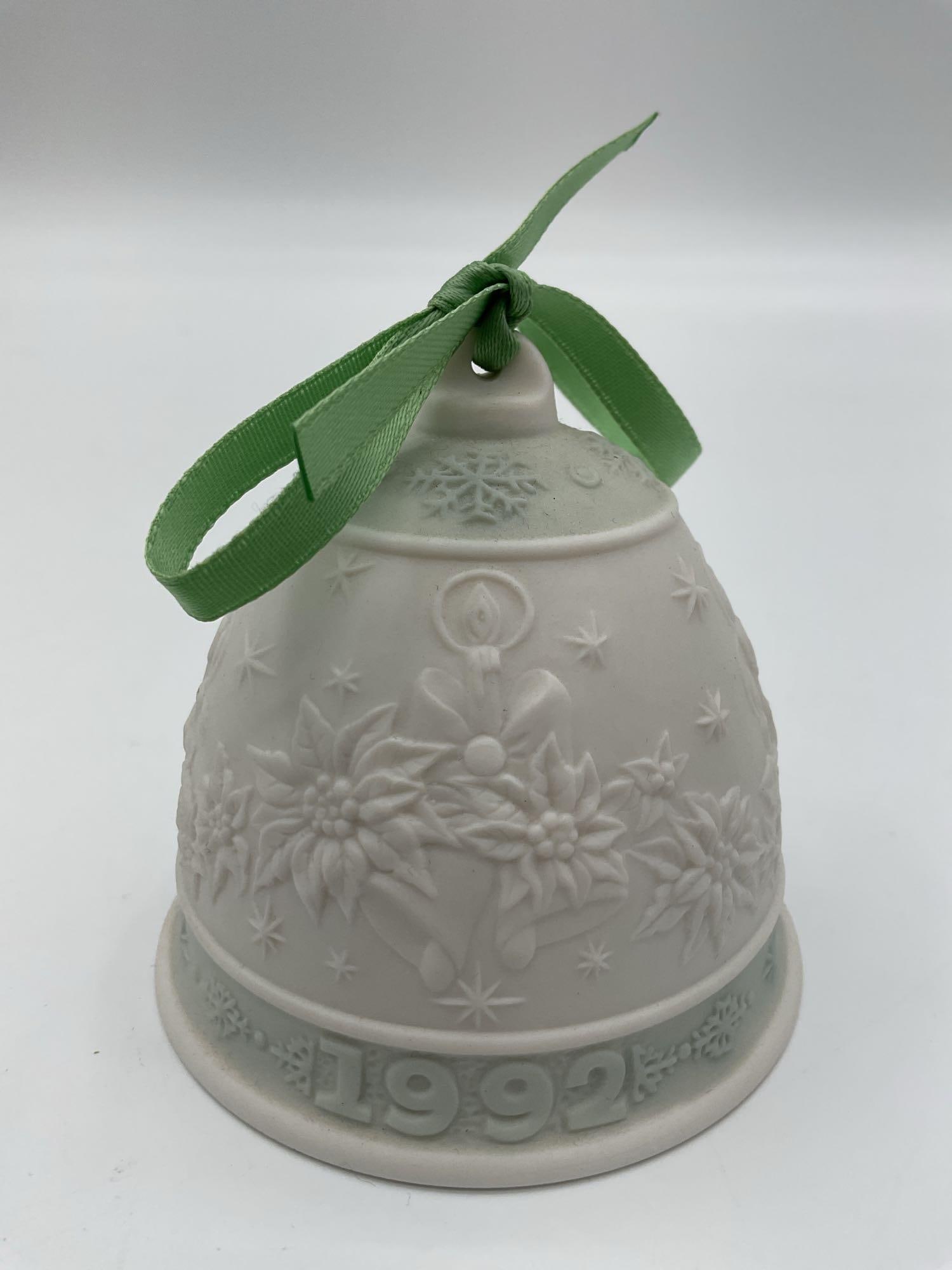 Lladro 1992 Christmas Bell Ornament:  A matte porcelain bell adorned with poinsettias, holly, and a central candle motif, topped with a green ribbon, and dated 1992. Dimensions: H3in Dia3in Medium: Unglazed (matte) porcelain