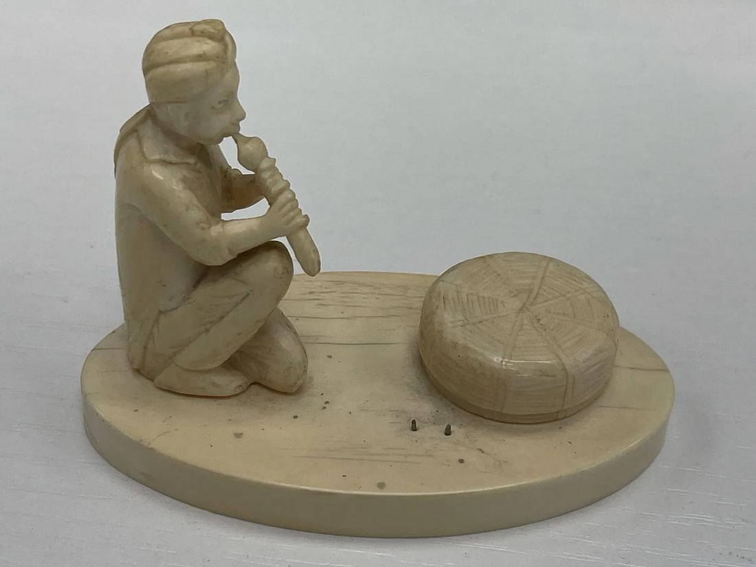 Snake Charmer Figurine: Hand-carved sculpture of a traditional snake charmer playing a flute, seated beside a basket on an oval base. The piece shows fine detail and craftsmanship, possibly of Indian origin.Dimensions: H2in