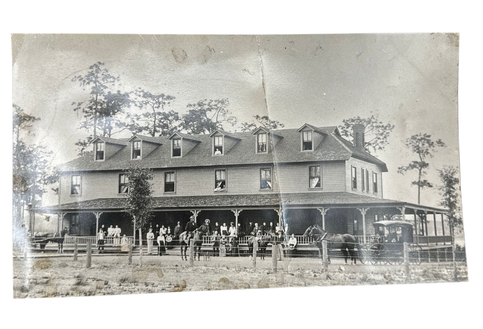 Early 20th Century Building Photograph: Black and white photograph depicting a large two-story wooden building with wraparound porch and dormer windows, surrounded by a fenced yard and tall trees. Several figures are visible standing along