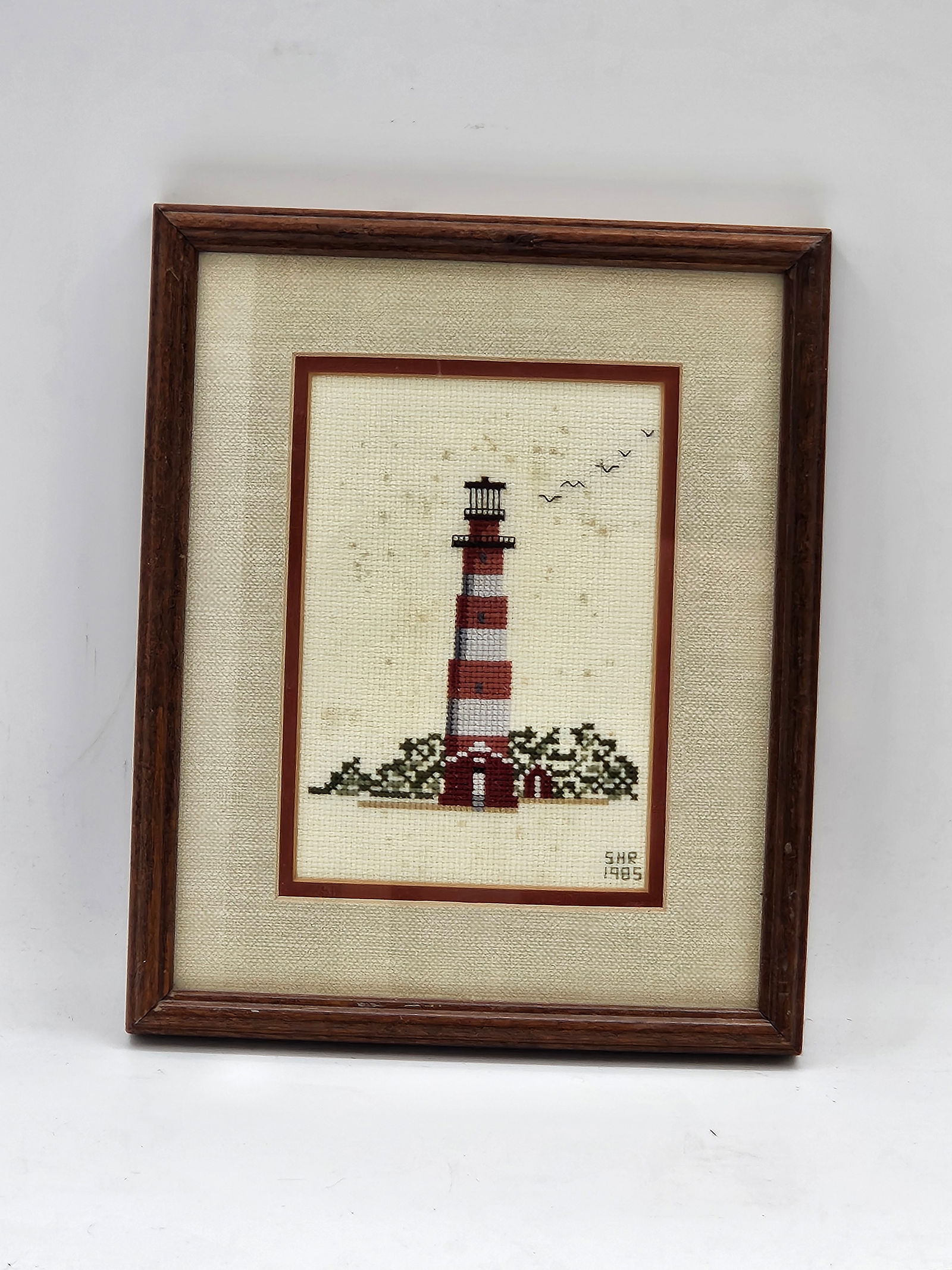 Framed Lighthouse Needlepoint: Hand-stitched needlepoint artwork depicting a classic red-and-white striped lighthouse surrounded by waves and flying birds. The simple design and warm tones evoke a charming coastal