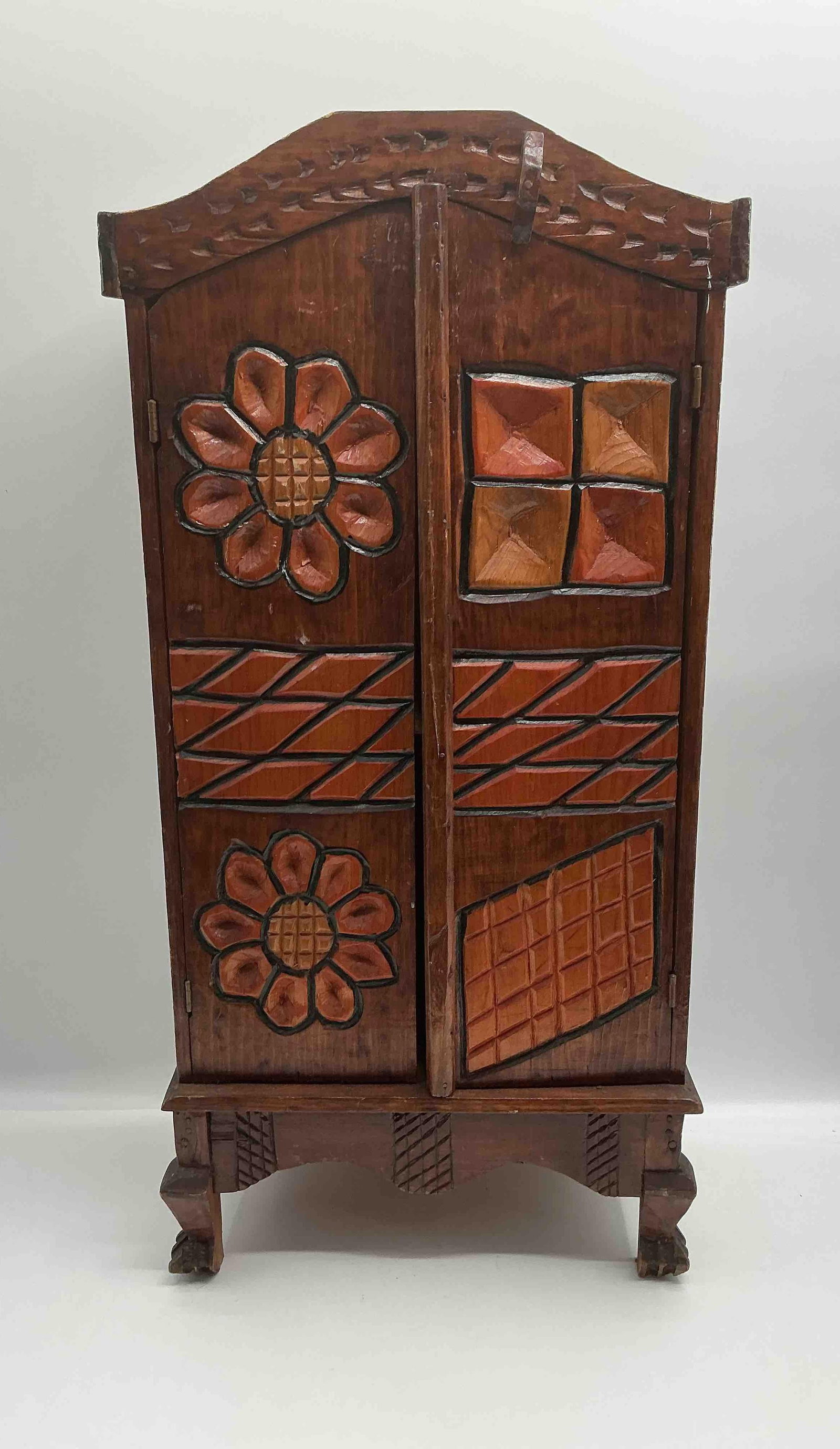Hand-Carved Folk Art Wooden Cabinet with Floral and Geometric Motifs: Small rustic cabinet featuring hand-carved and painted floral and geometric designs in warm earth tones. Raised on four sturdy legs with decorative carvings, this piece has a charming folk-art appeal