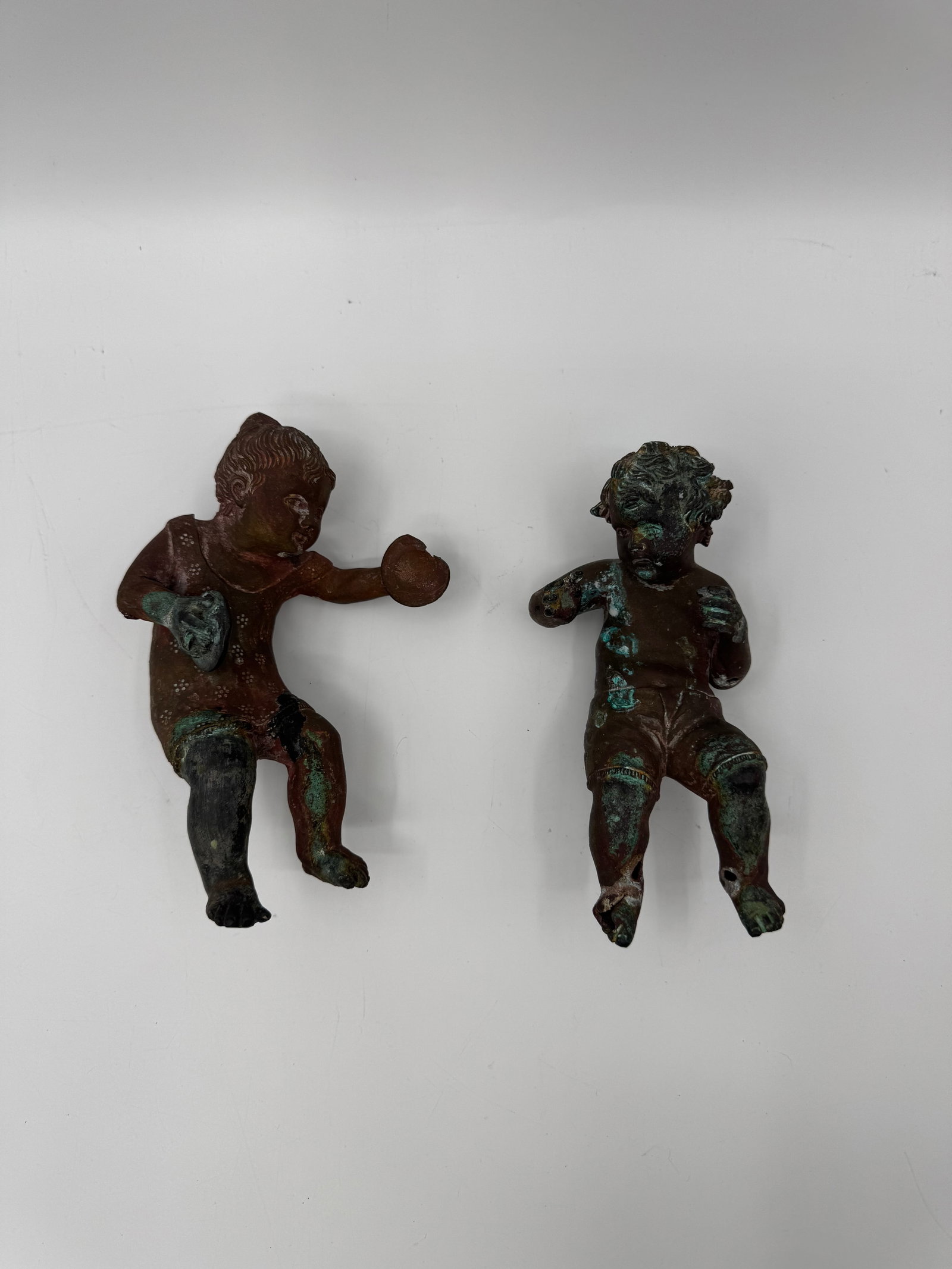 Pair of Antique Bronze Cherub Figures: Charming pair of small antique bronze cherub figures with aged patina. Each cherub is depicted in motion holding an object, with hollow backs suggesting they may have been decorative mounts or