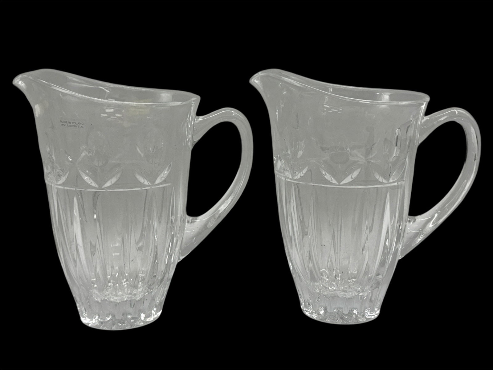 Pair of Cut Glass Pitchers: A pair of clear cut glass pitchers featuring flared rims, molded vertical paneling, and decorative crosshatch detailing near the upper body. Each pitcher includes a curved handle and shaped spout, com