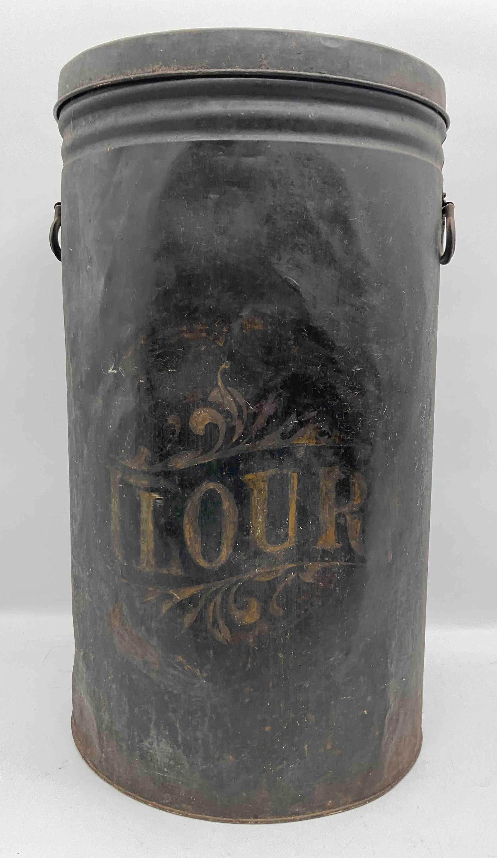 Antique Flour Storage Tin: Large black painted tin container with faded decorative lettering reading “Flour.” Features side handles and removable lid. Bottom is missing. Likely early to mid-20th century kitchen stor