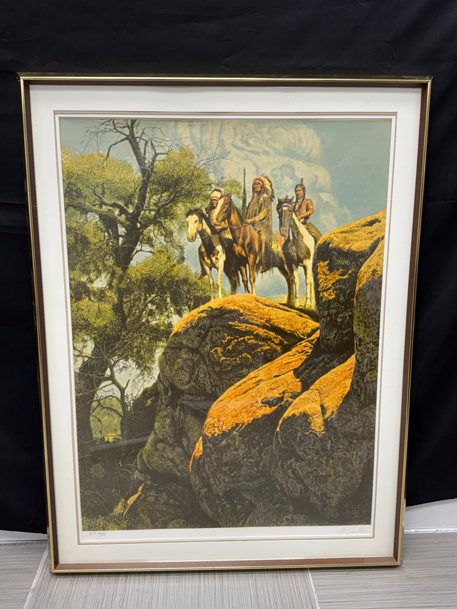 Framed Print of Native Americans on Horseback by Mort Kunstler: Framed print depicting Native Americans on horseback riding through a rocky landscape with trees. The print is framed in a gold-tone metal frame. The image is detailed and colorful.