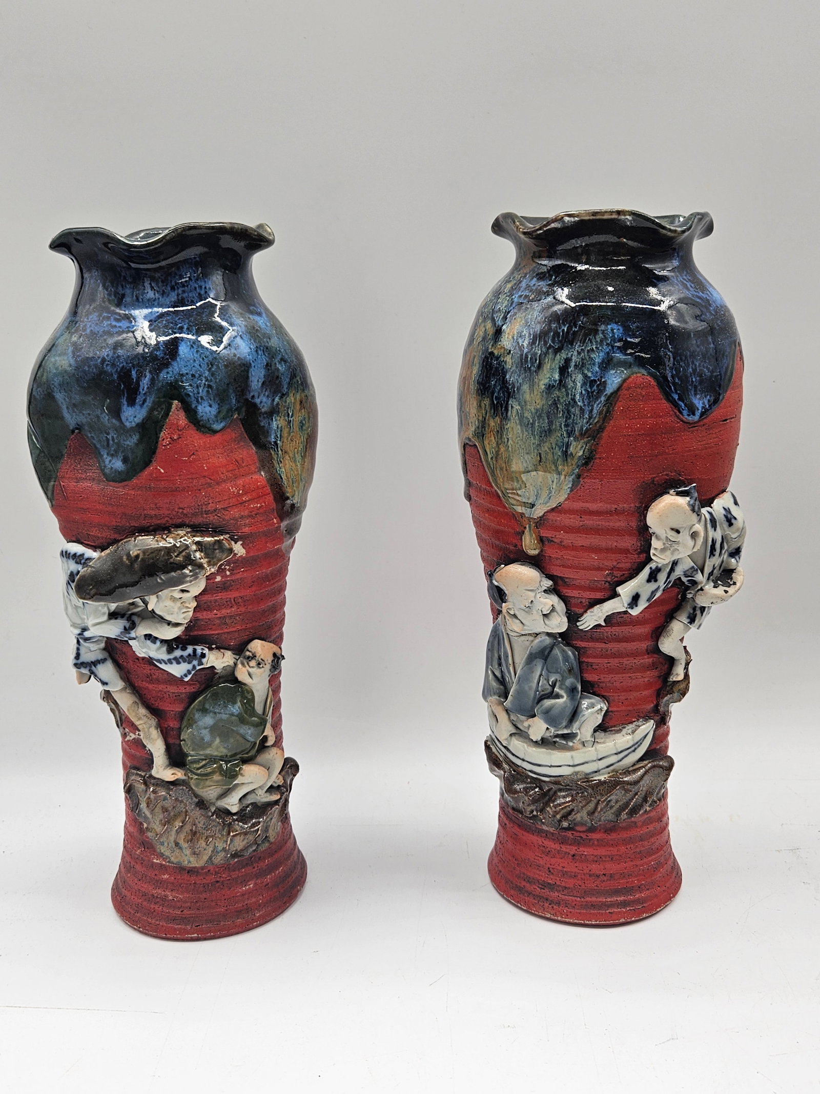 Pair of Japanese Satsuma Figural Vases:  Decorative pair of tall vases featuring raised figural scenes of traditional Japanese figures in relief against a red ground with blue glazed rims. The textured design and expressive characters