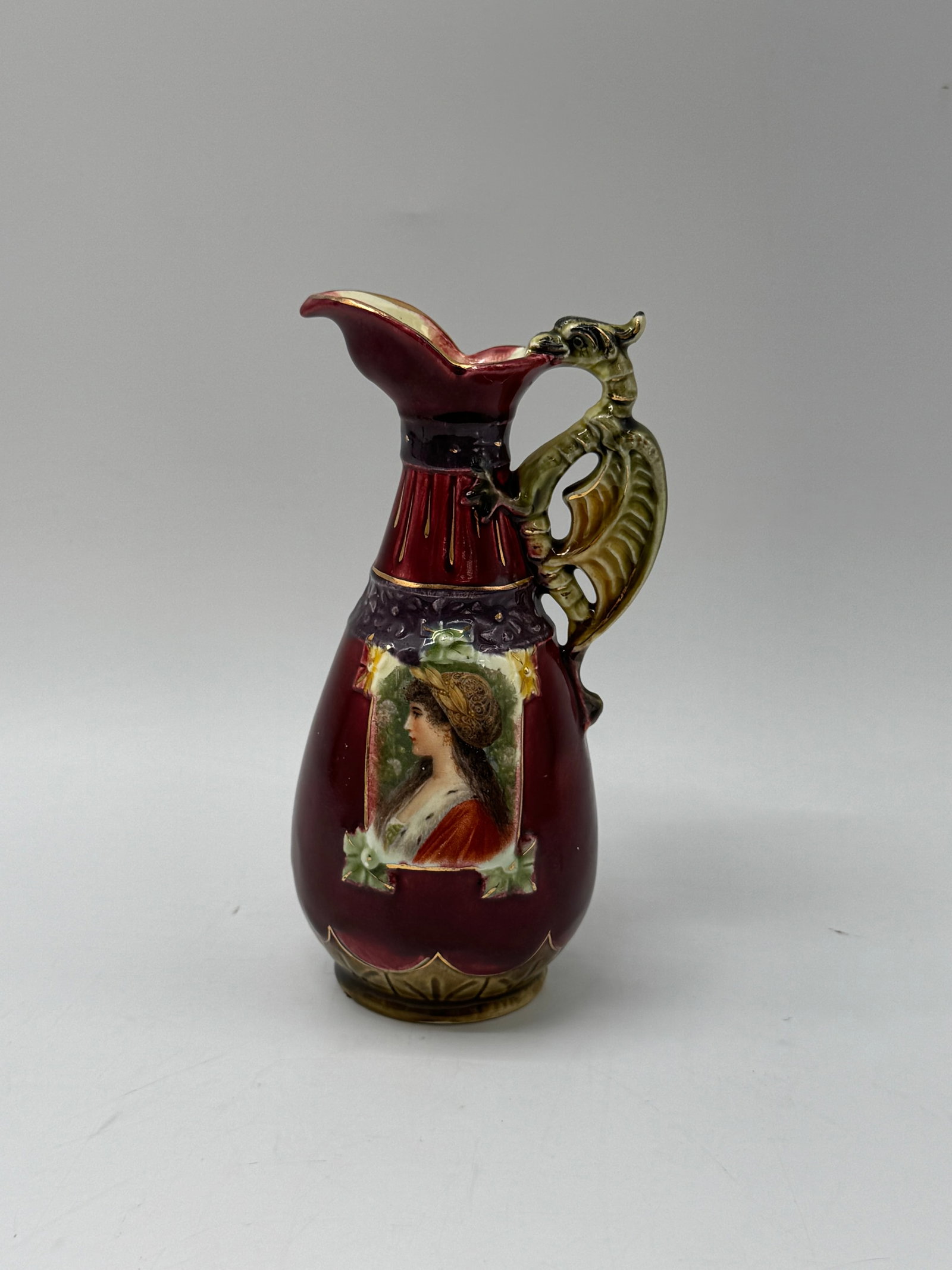 Majolica Style Ewer with Portrait and Dragon Handle: A decorative majolica style ewer featuring a portrait medallion and a dragon-shaped handle. The ewer has a rich burgundy glaze with gilt accents. It is unmarked. Measures approximately 7 inches tall a